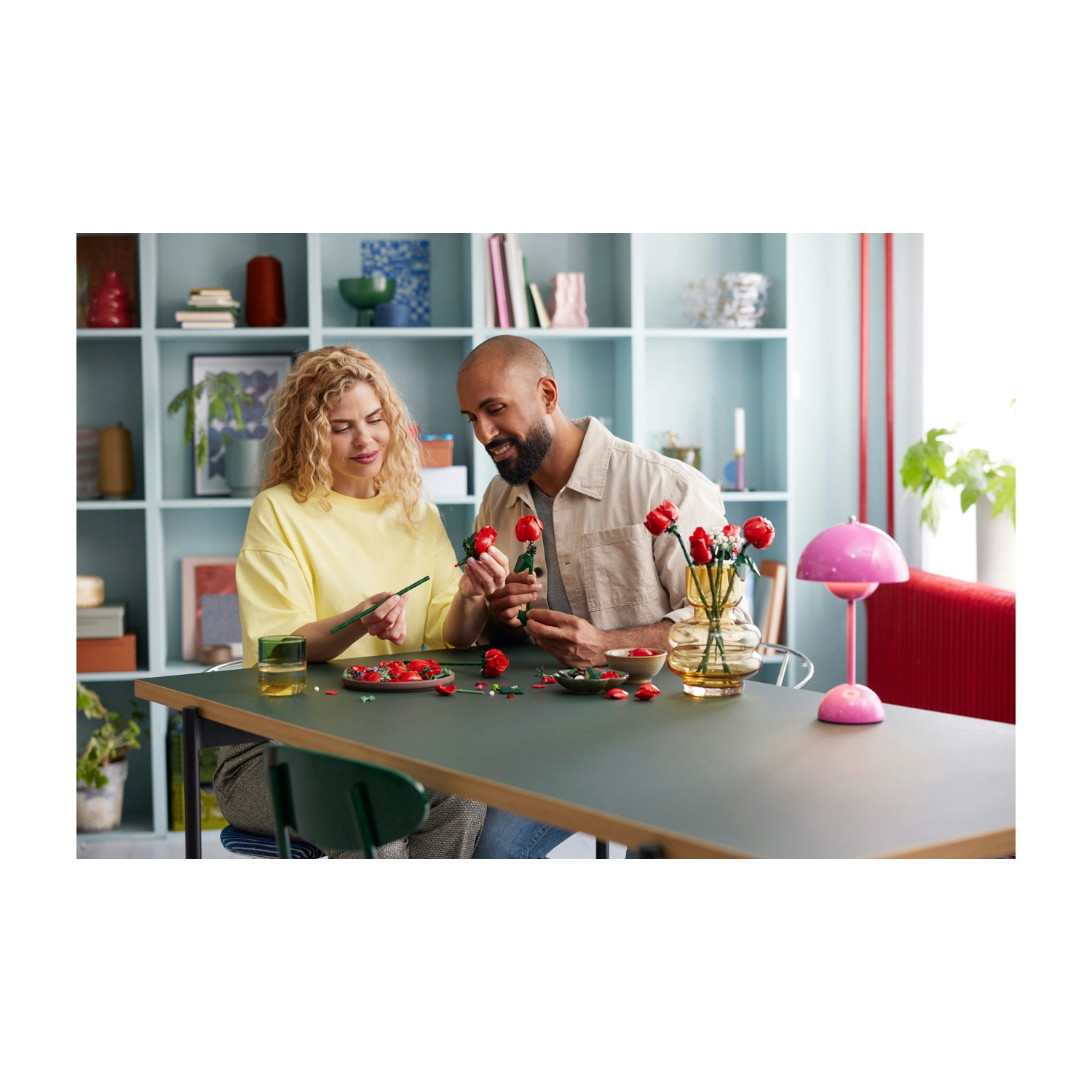 Una coppia assembla rose Lego a un tavolo. Un bicchiere d'acqua, petali di rosa e una lampada rosa sono sul tavolo. Un vaso con rose sullo sfondo.