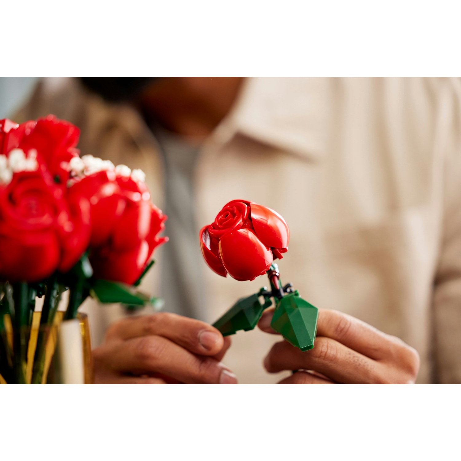 Una persona tiene in mano una rosa Lego. Un altro mazzo di rose Lego in un vaso è a sinistra. Lo sfondo è sfocato.