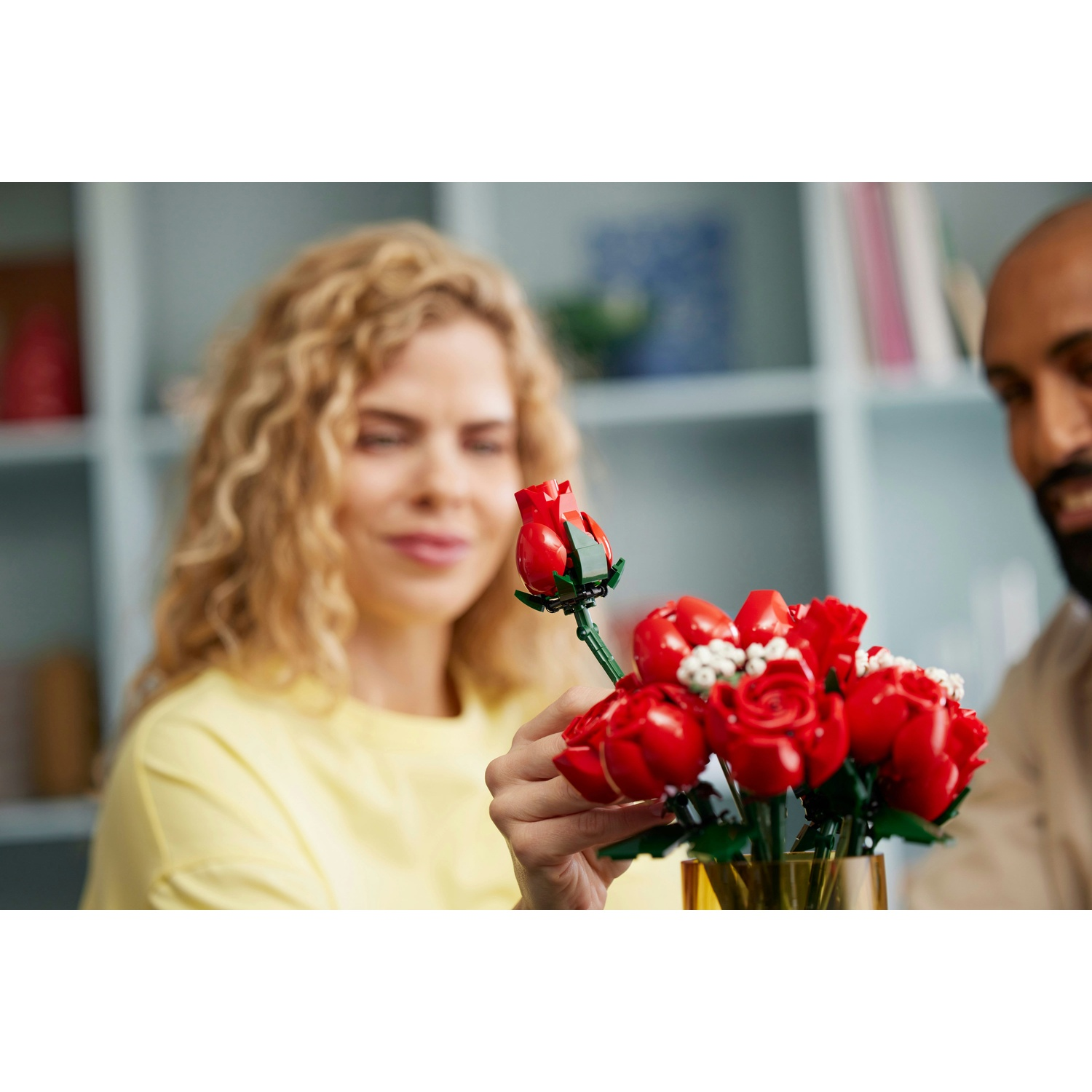 Una donna tiene una rosa rossa da un bouquet Lego. Un uomo è accanto a lei. Il bouquet è in un vaso giallo.