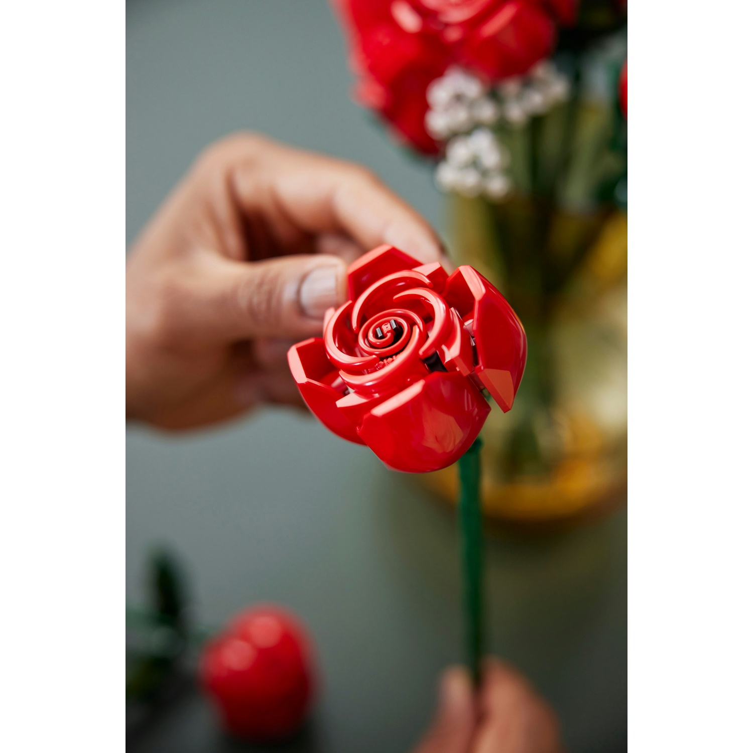 Una mano tiene in mano una rosa di plastica rossa. Un mazzo di rose simili è sullo sfondo. La rosa ha un gambo verde.