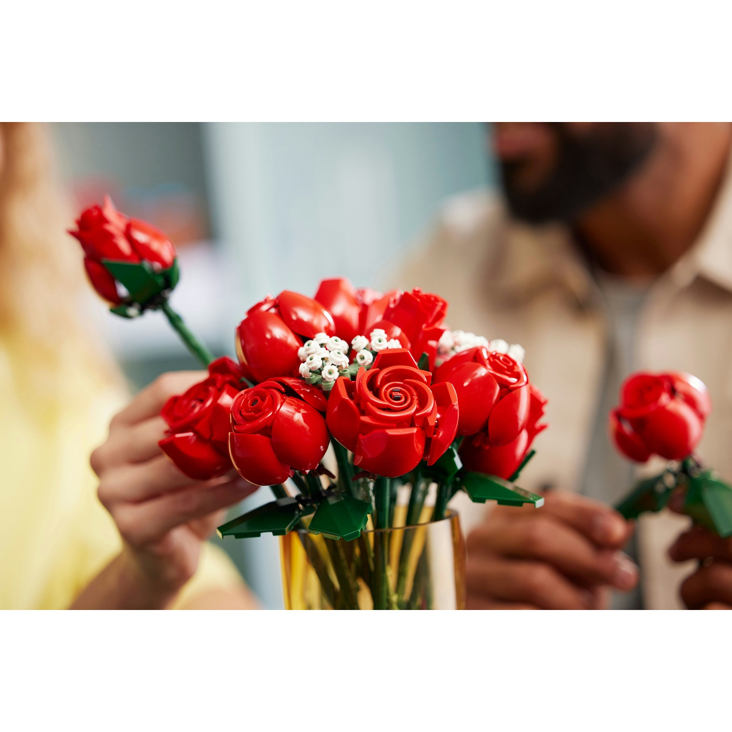 Un bouquet di rose rosse Lego in un vaso d'oro. Due persone stanno sistemando i fiori. Lo sfondo è sfocato.