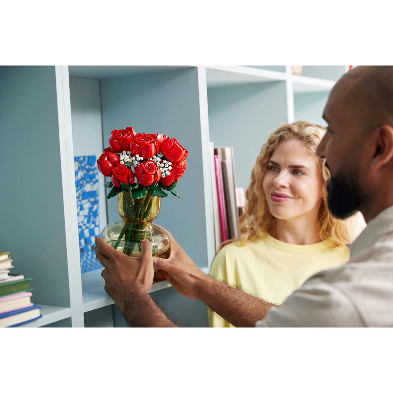 Un uomo tiene un vaso di rose rosse, osservato da una donna. Le librerie sono sullo sfondo. Il vaso è su uno scaffale.