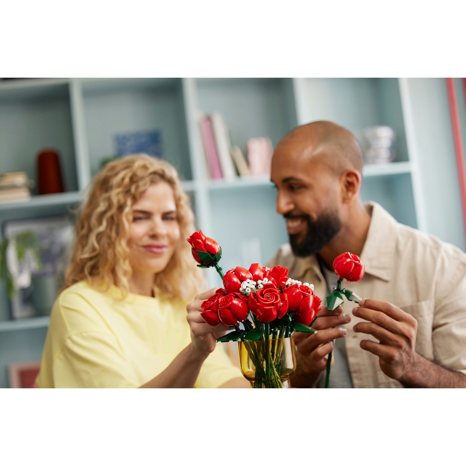 Un uomo e una donna sistemano dei bouquet di rose rosse. Sorridono. Un vaso giallo contiene delle rose. Scaffali sullo sfondo.