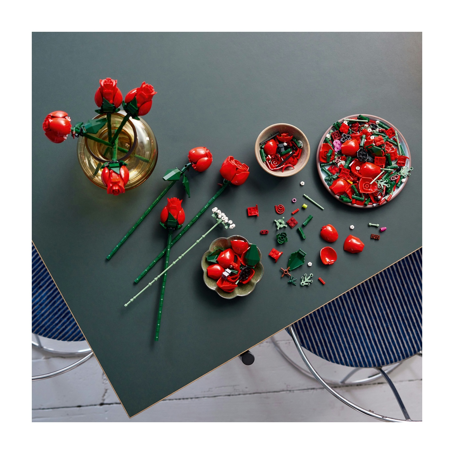 Set Lego di rose rosse disposto su un tavolo verde scuro. Include un vaso, ciotole con pezzi e fiori assemblati. Due sedie visibili.