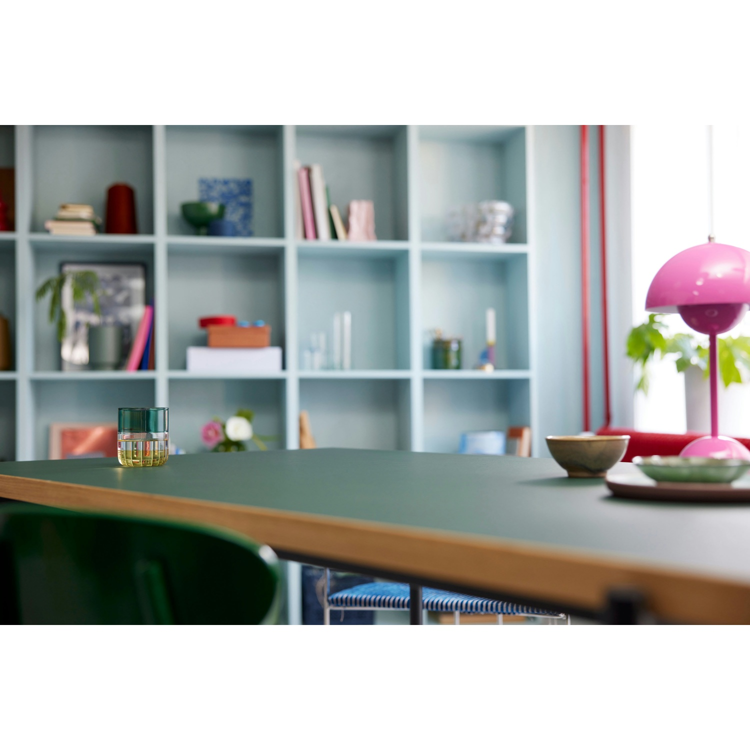 Un bicchiere verde è su un tavolo verde. Una lampada rosa e delle ciotole sono sul tavolo. Una libreria sullo sfondo.