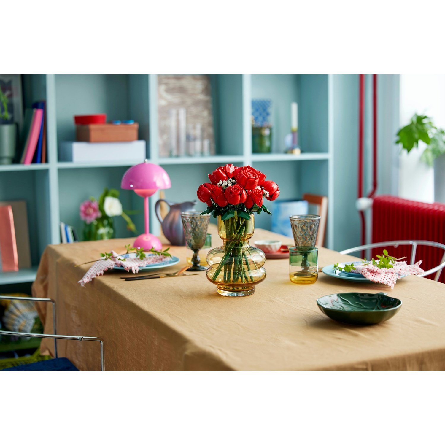 Una tavola apparecchiata con rose rosse in un vaso. Lampada rosa, bicchieri, piatti e una ciotola verde sono presenti, su una tovaglia marrone.
