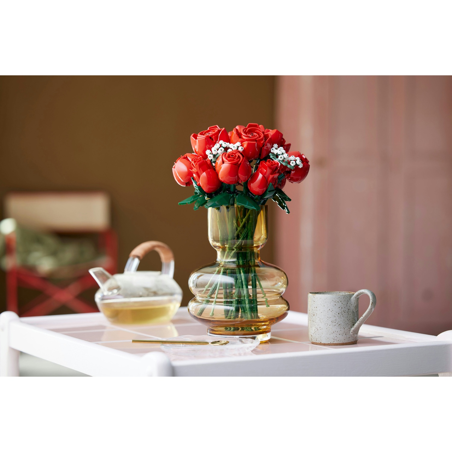 Un mazzo di rose rosse in un vaso di vetro è su un tavolo bianco, vicino a una tazza e una teiera. Lo sfondo è sfocato.