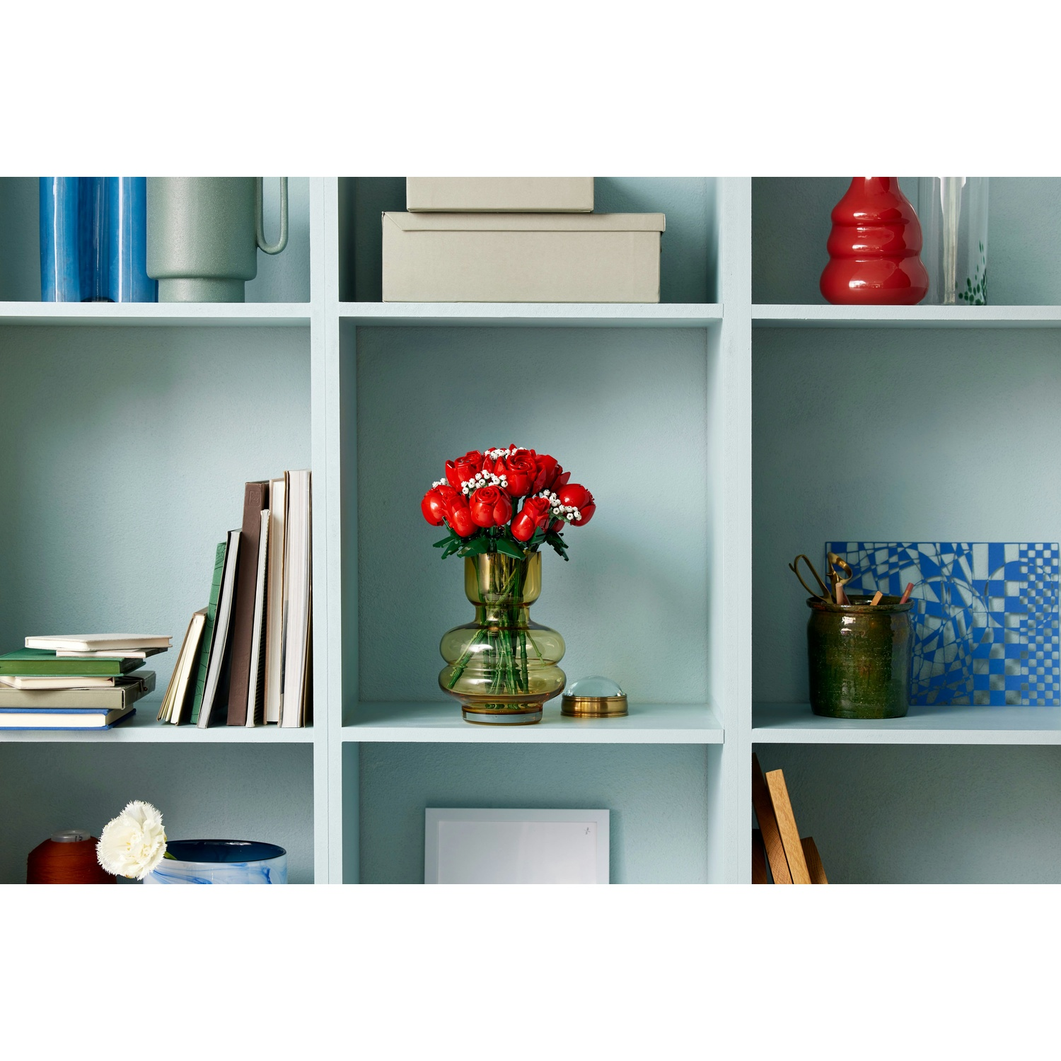 Una mensola con vari oggetti: libri, scatole, vasi con fiori e oggetti decorativi. I colori includono rosso, verde, blu e bianco.
