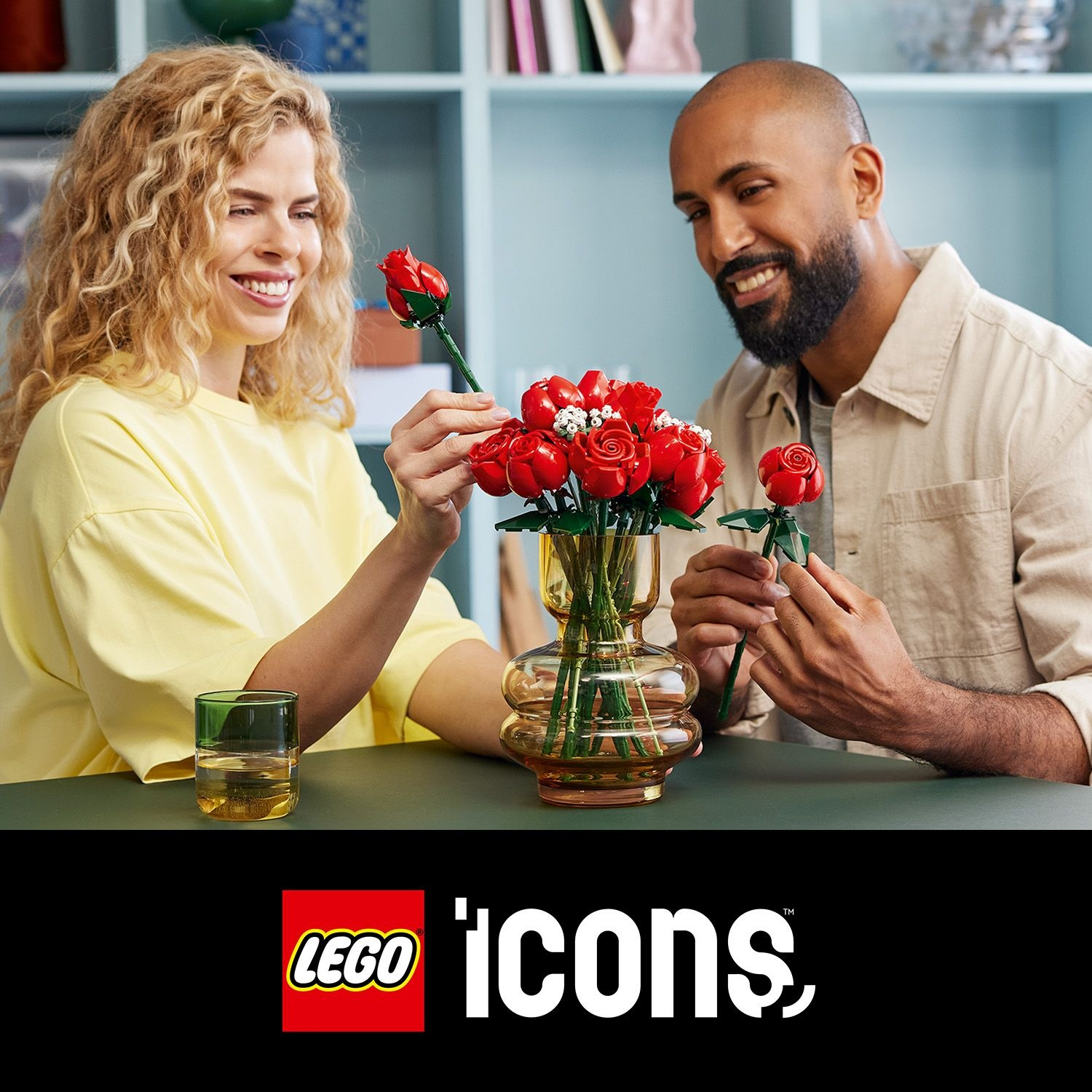 Una coppia sorride, disponendo rose Lego in un vaso. Un bicchiere è sul tavolo. Il logo Lego e 'icons' in basso.
