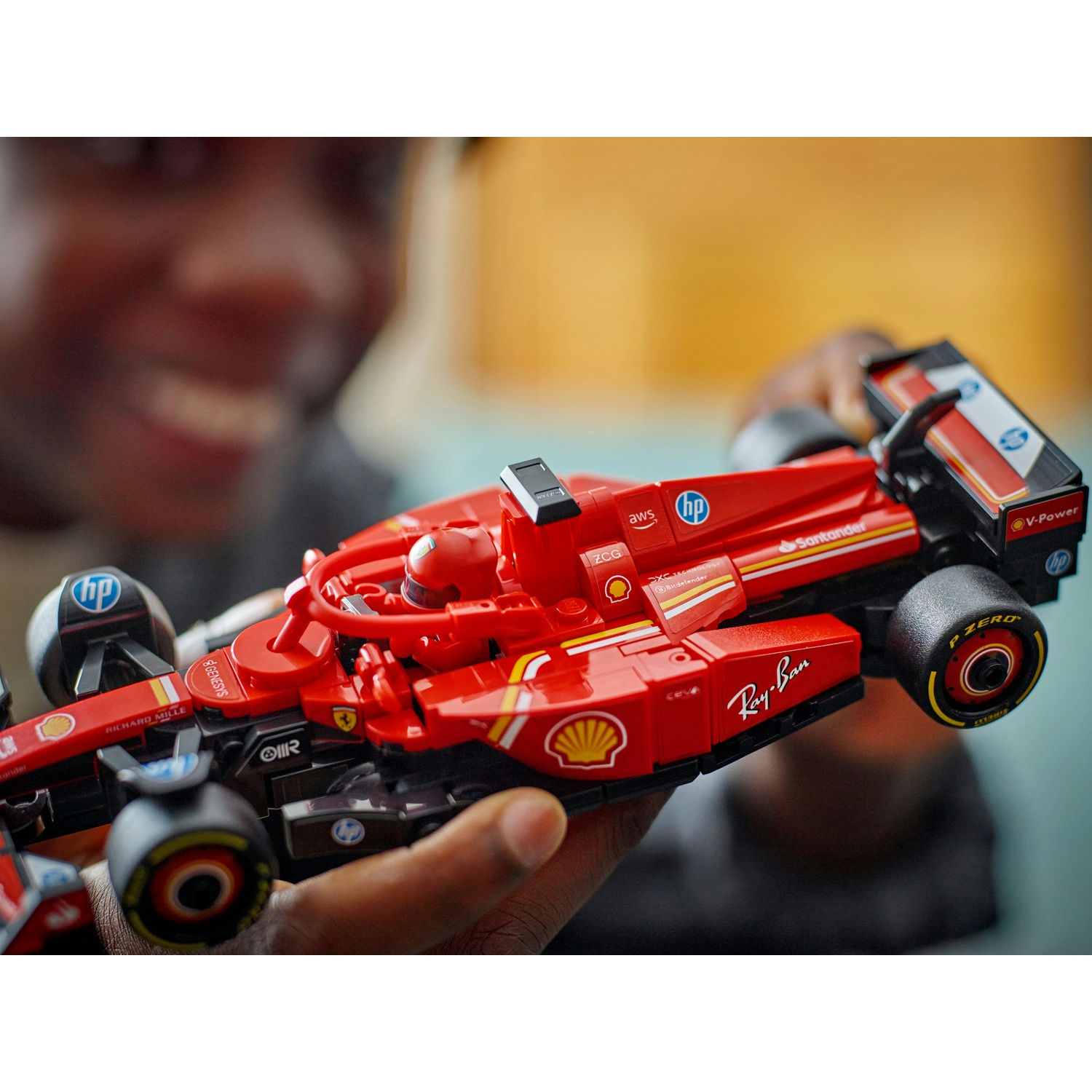 Una persona tiene in mano una macchina di F1 Lego rossa. I loghi includono HP, Santander, Shell e Ray-Ban. L'auto ha pneumatici neri e un pilota.