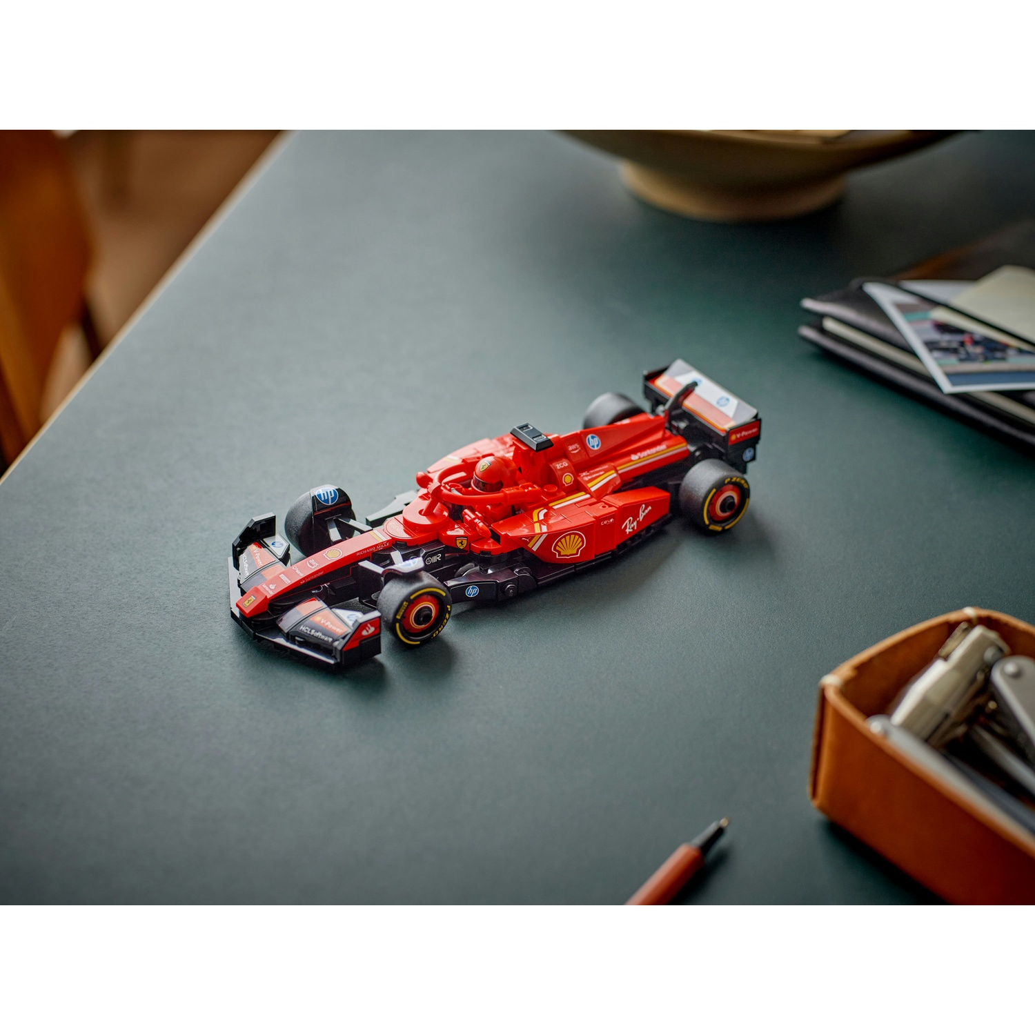 Un modello di auto da corsa Ferrari rossa è su una superficie verde scuro. Una penna, un vassoio con oggetti e dei fogli sono anche sul tavolo.