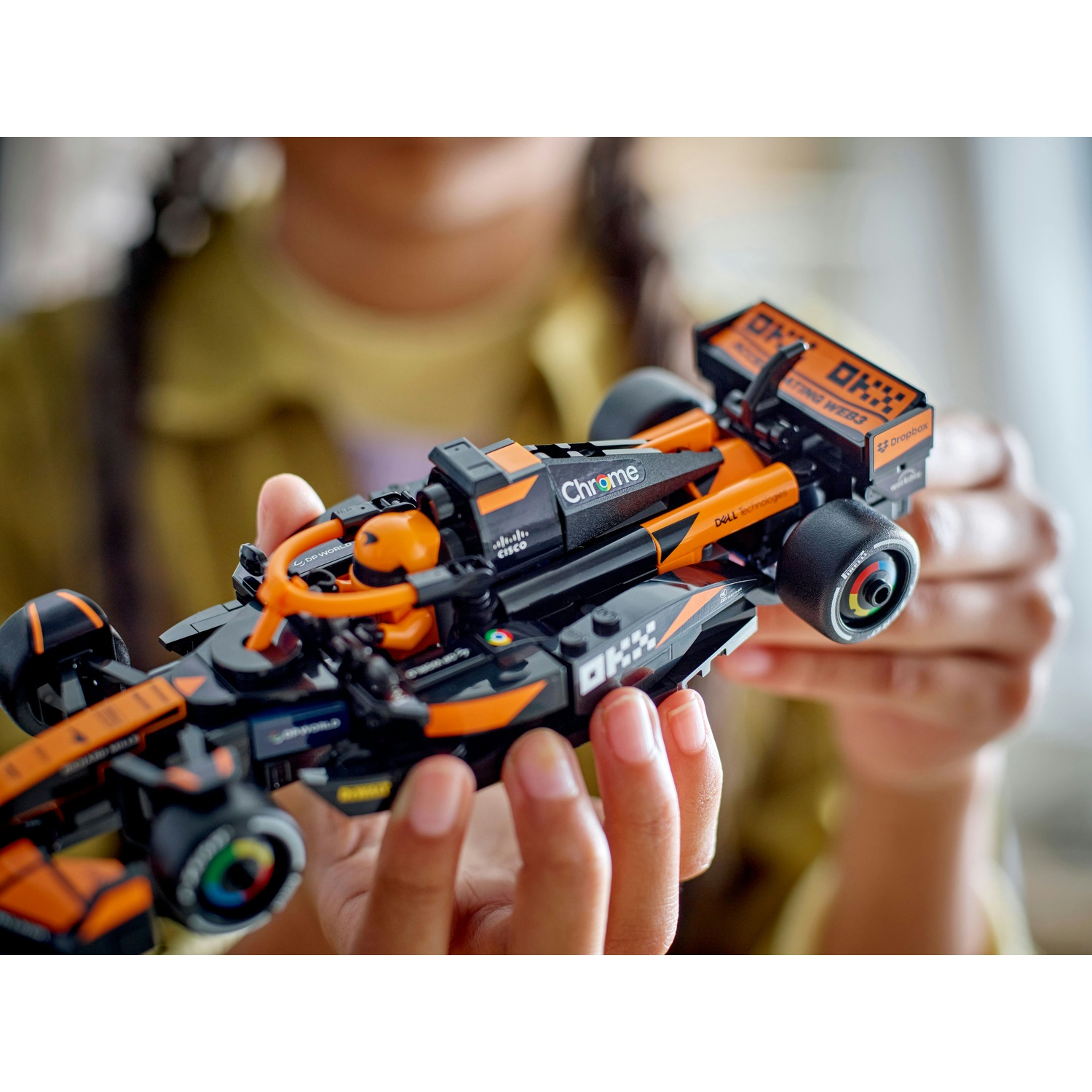 Una persona tiene in mano una macchina da corsa Lego. L'auto è nera e arancione con loghi degli sponsor. C'è una figura di pilota. Lo sfondo è sfocato.