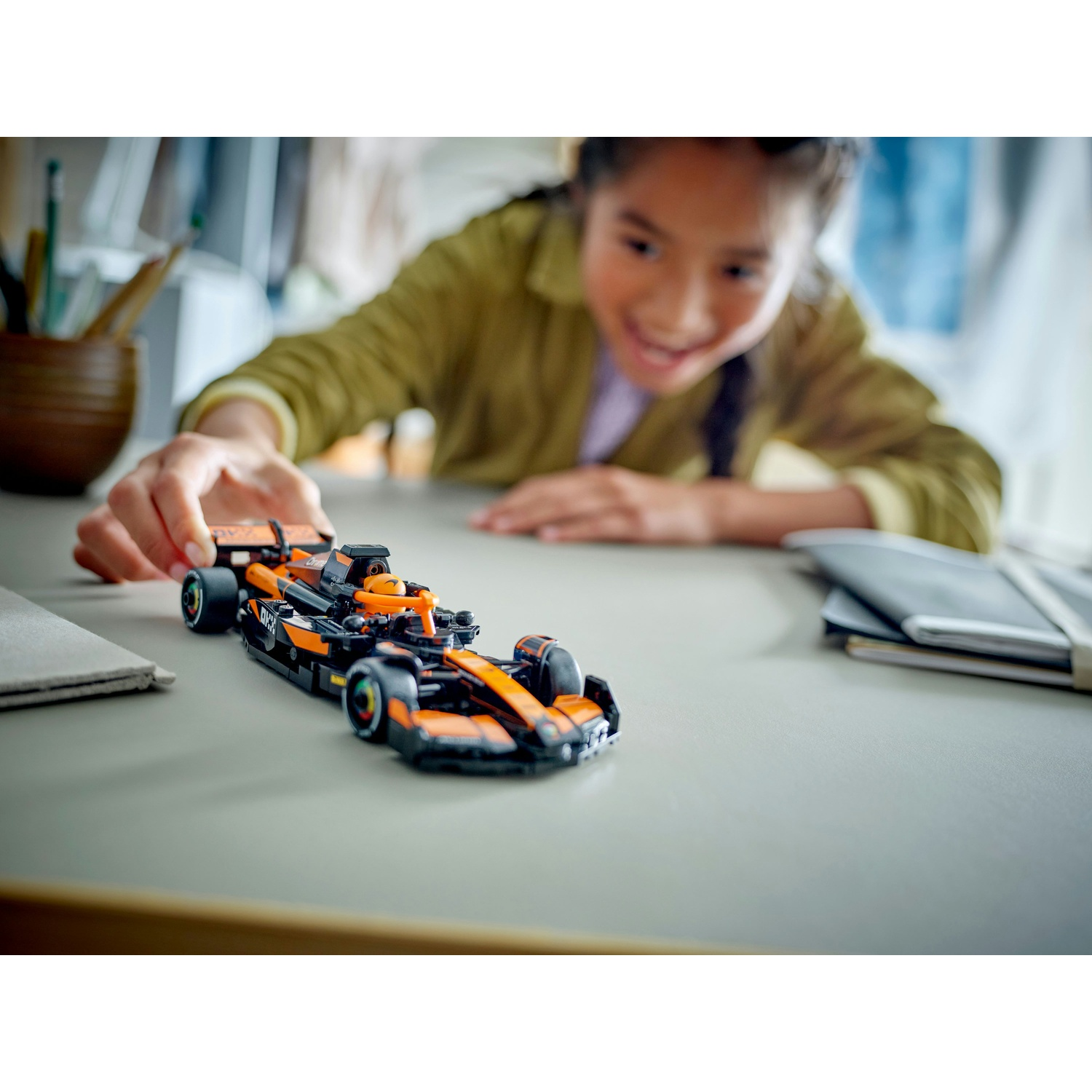 Una ragazza sorride, allungando la mano verso una macchina da corsa Lego su un tavolo. L'auto è nera e arancione. Altri oggetti sono sfocati sullo sfondo.