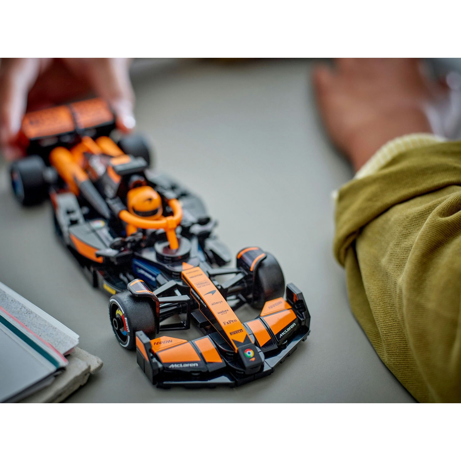 Le mani di una persona tengono un'auto giocattolo McLaren F1 arancione e nera. L'auto è su una superficie grigia con carta e un braccio.