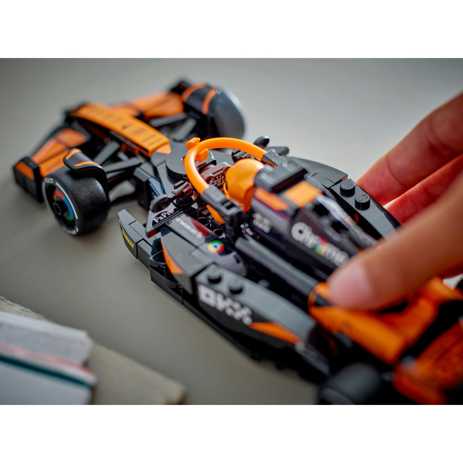 Una mano tiene in mano una macchina da corsa Lego nera e arancione su una superficie grigia. L'auto ha dettagli e adesivi visibili, con un volante.