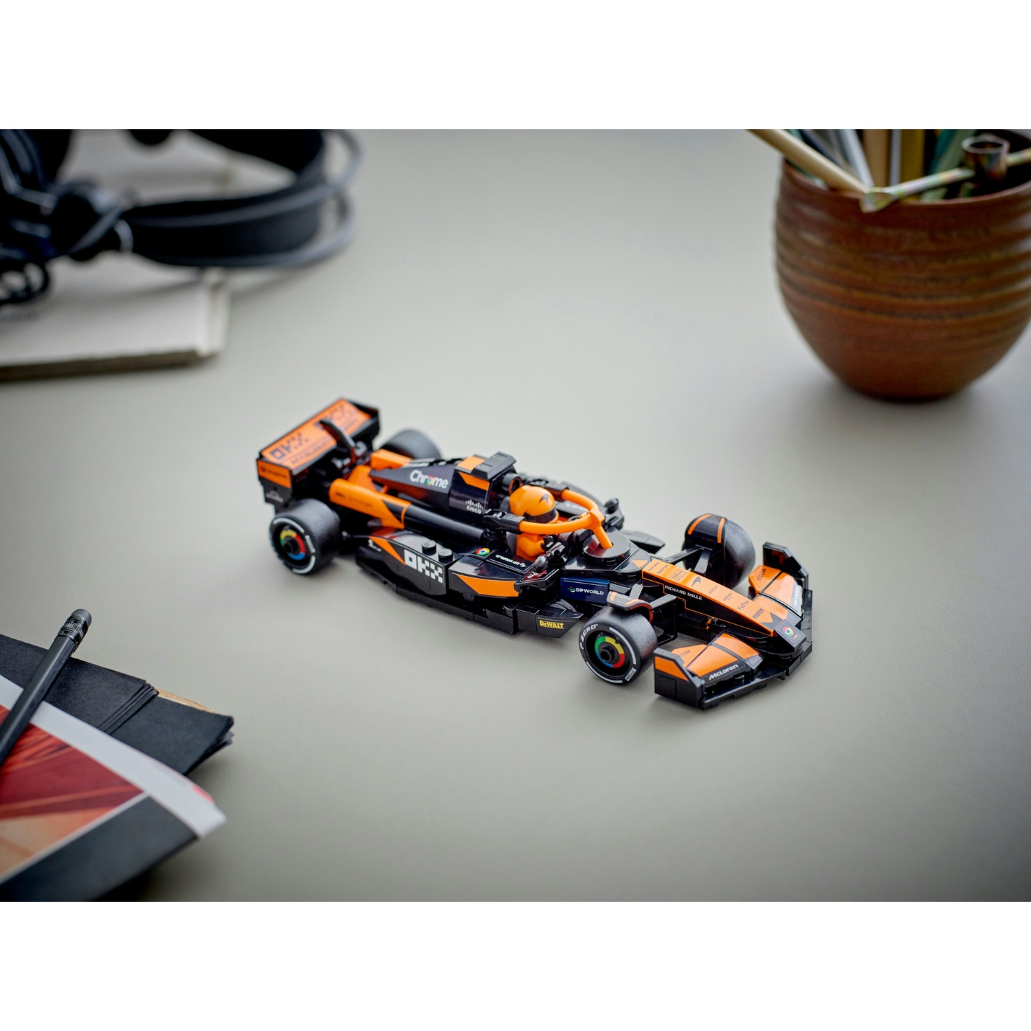 Un'auto giocattolo di Formula 1, arancione e nera, è su una superficie grigia. È circondata da forniture per ufficio: matite, blocco note e cuffie.