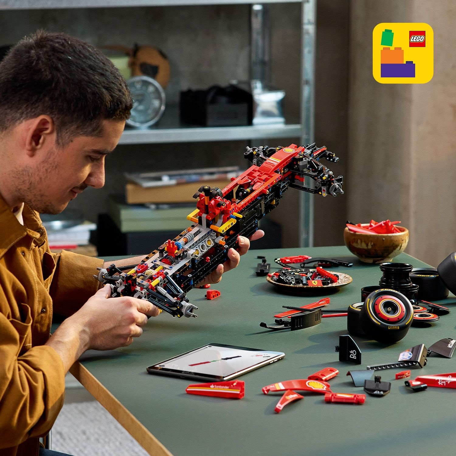 Un uomo assembla un'auto da corsa Lego su un tavolo verde. Varie parti sono sparse. Un tablet mostra le istruzioni, il logo Lego è visibile.