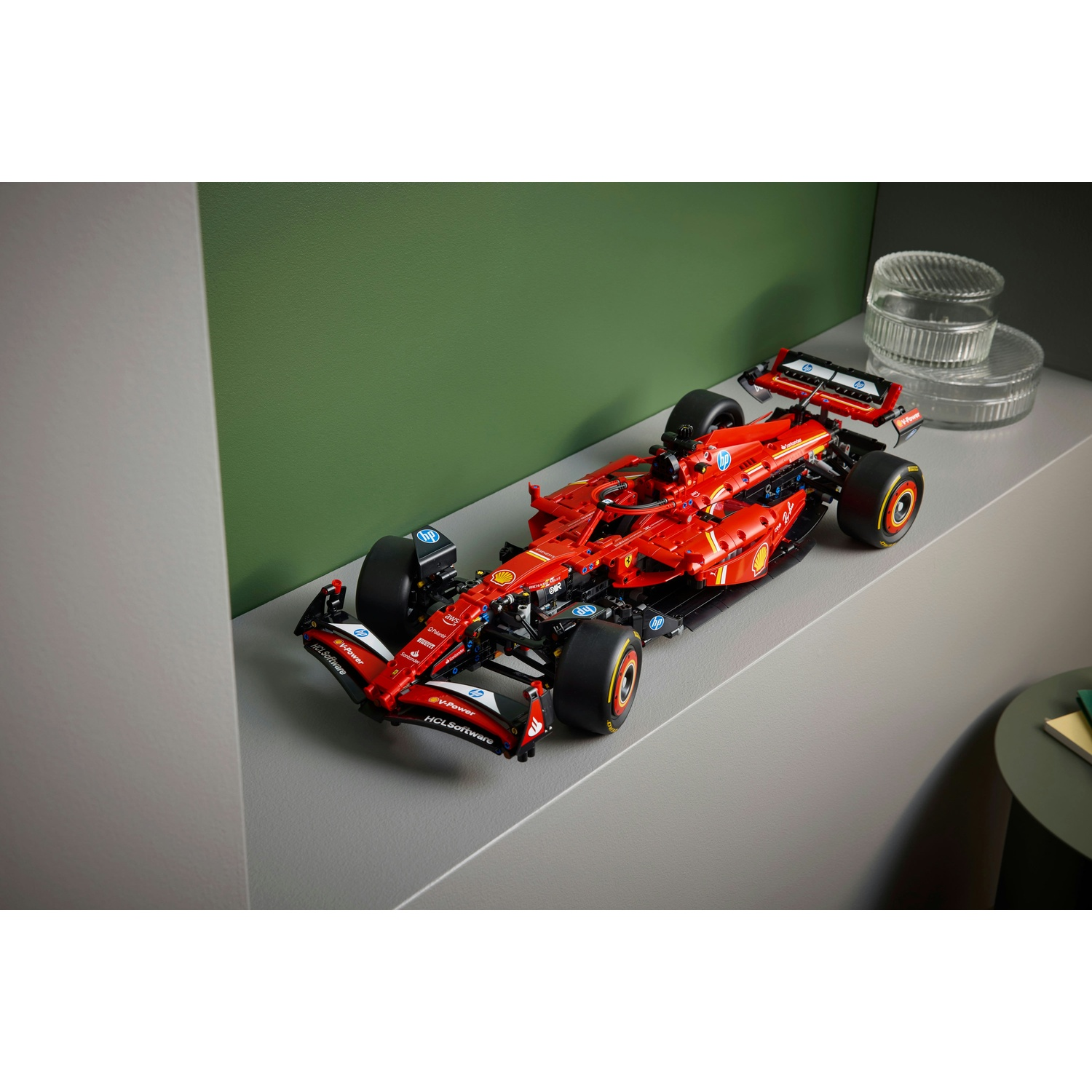 Un modello di auto F1 Lego rosso su una superficie grigia. Parete verde sullo sfondo, con contenitori di vetro su uno scaffale. Tavolo a destra.