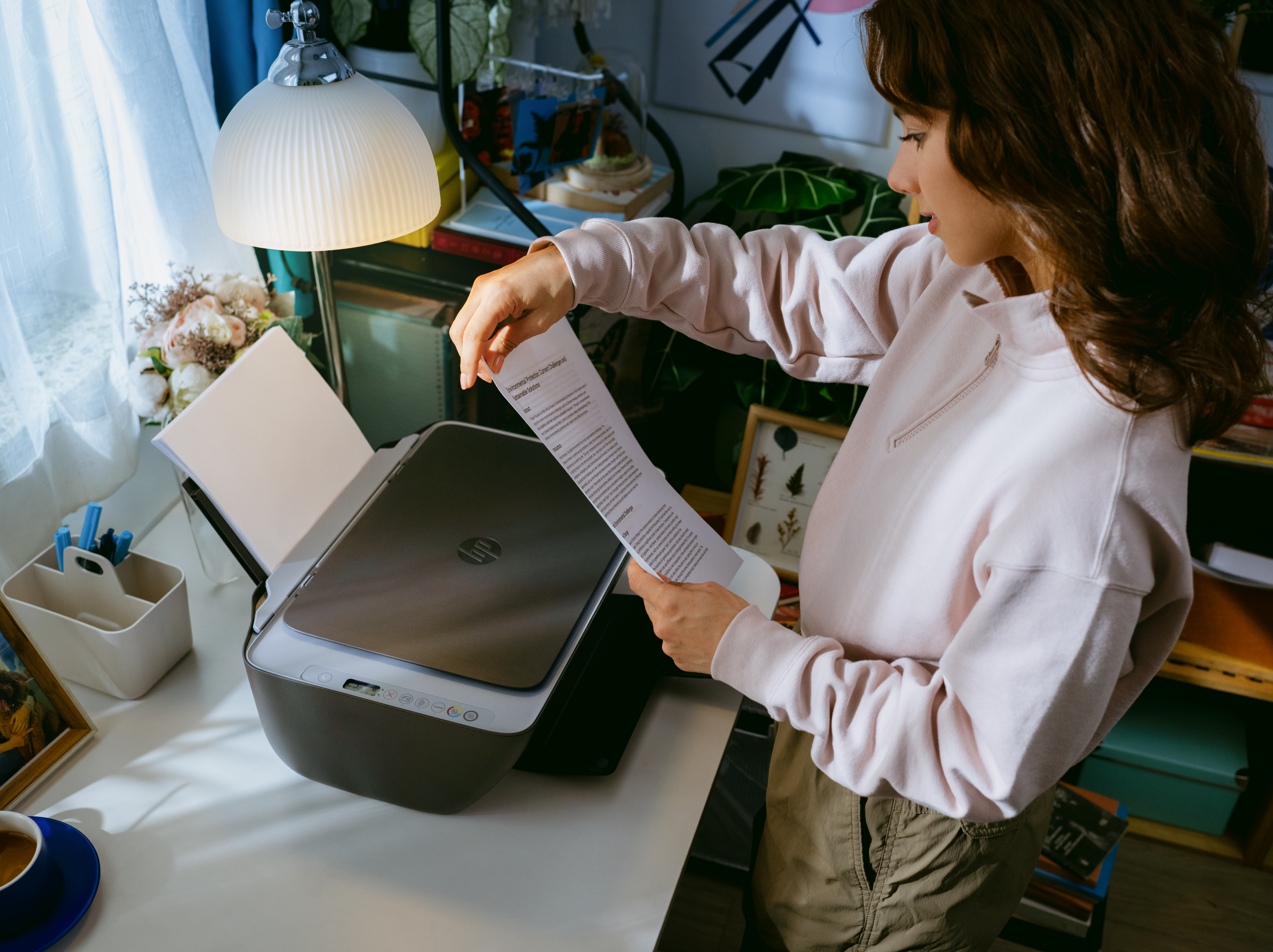 Eine Frau druckt ein Papier mit einem HP-Drucker auf einem weißen Schreibtisch. Eine Lampe, Blumen und Bücher sind im Hintergrund.