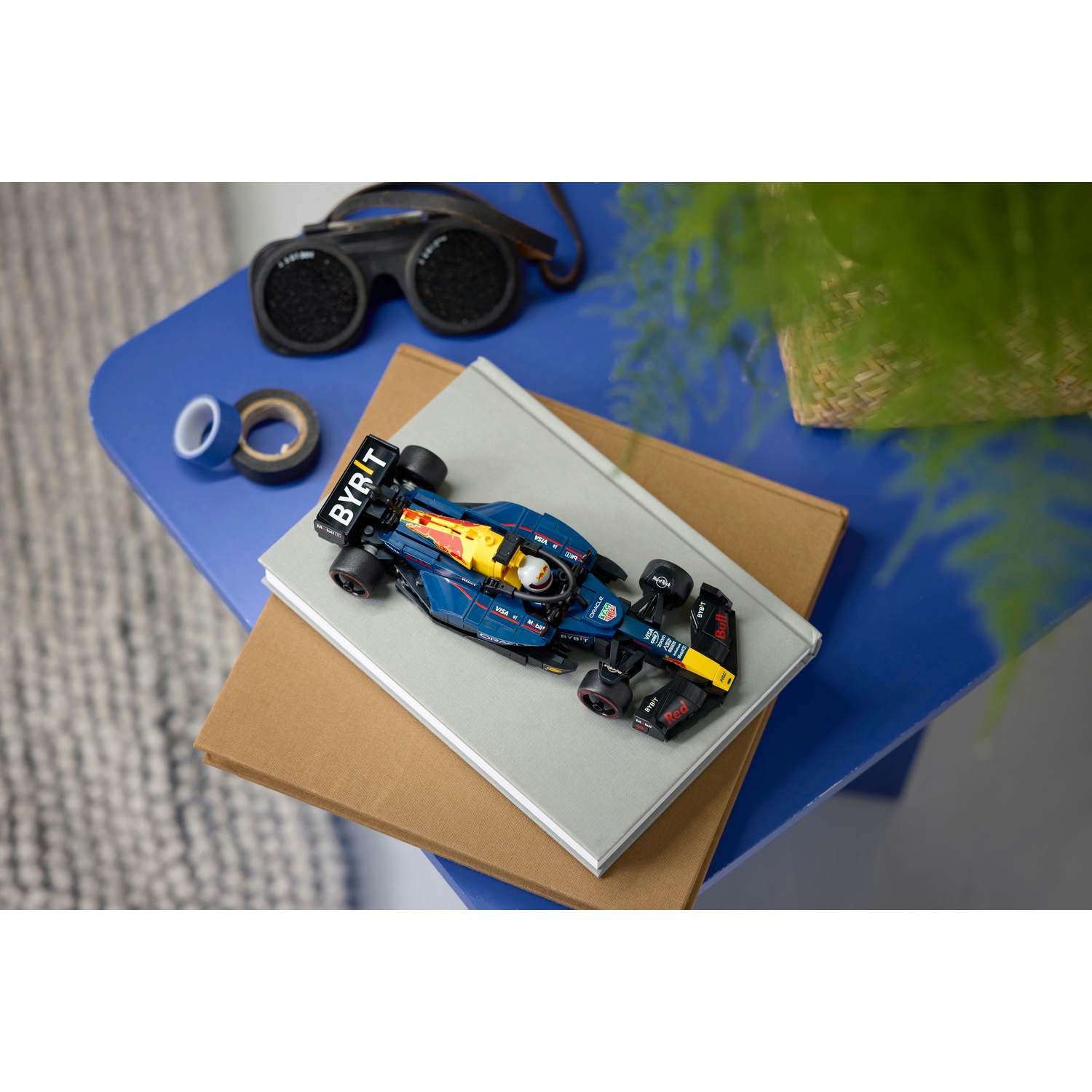 Un'auto giocattolo di Formula 1 su libri, nastro adesivo e occhiali su un tavolo blu, con una pianta sullo sfondo.