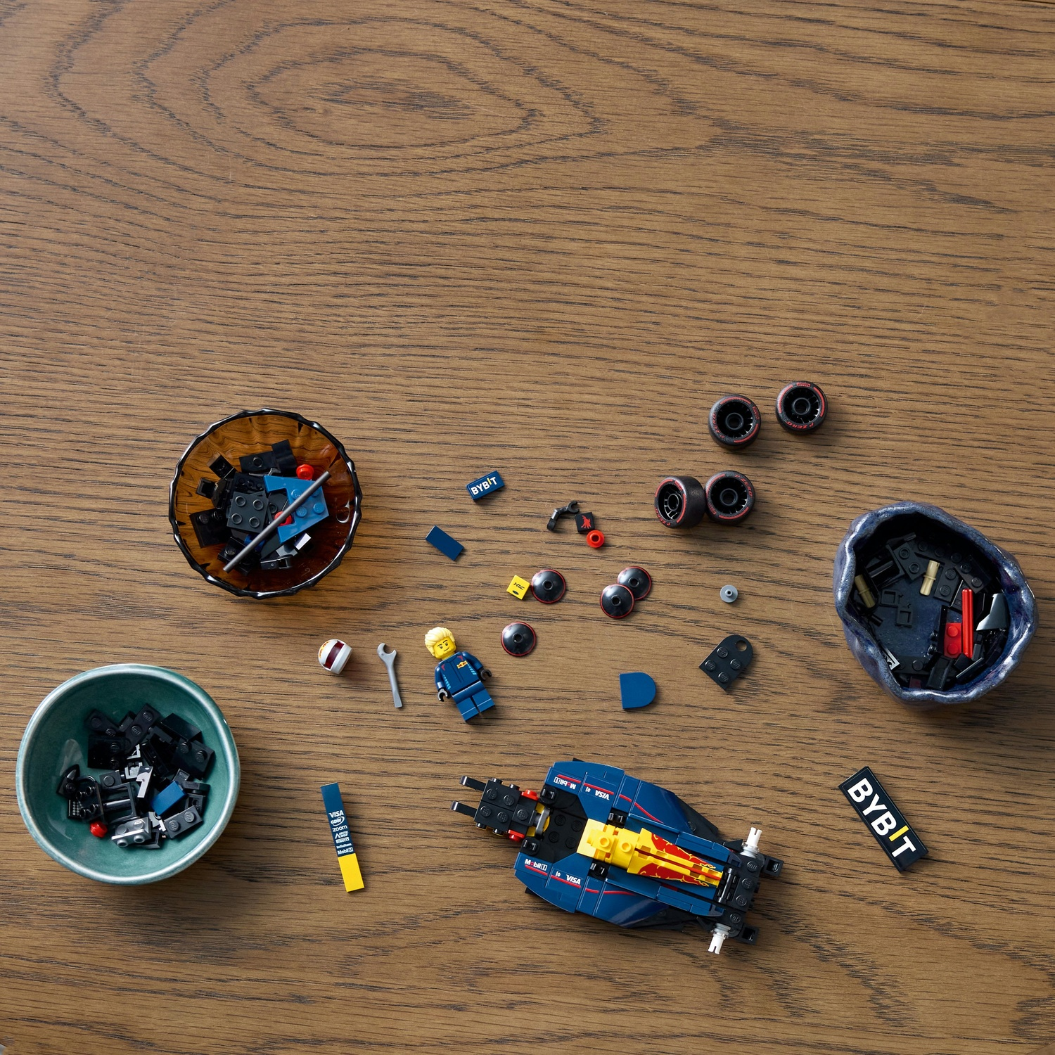 Set di auto da corsa Lego con pezzi sparsi, ruote, minifigure e ciotole su un tavolo di legno. Include il marchio dello sponsor.