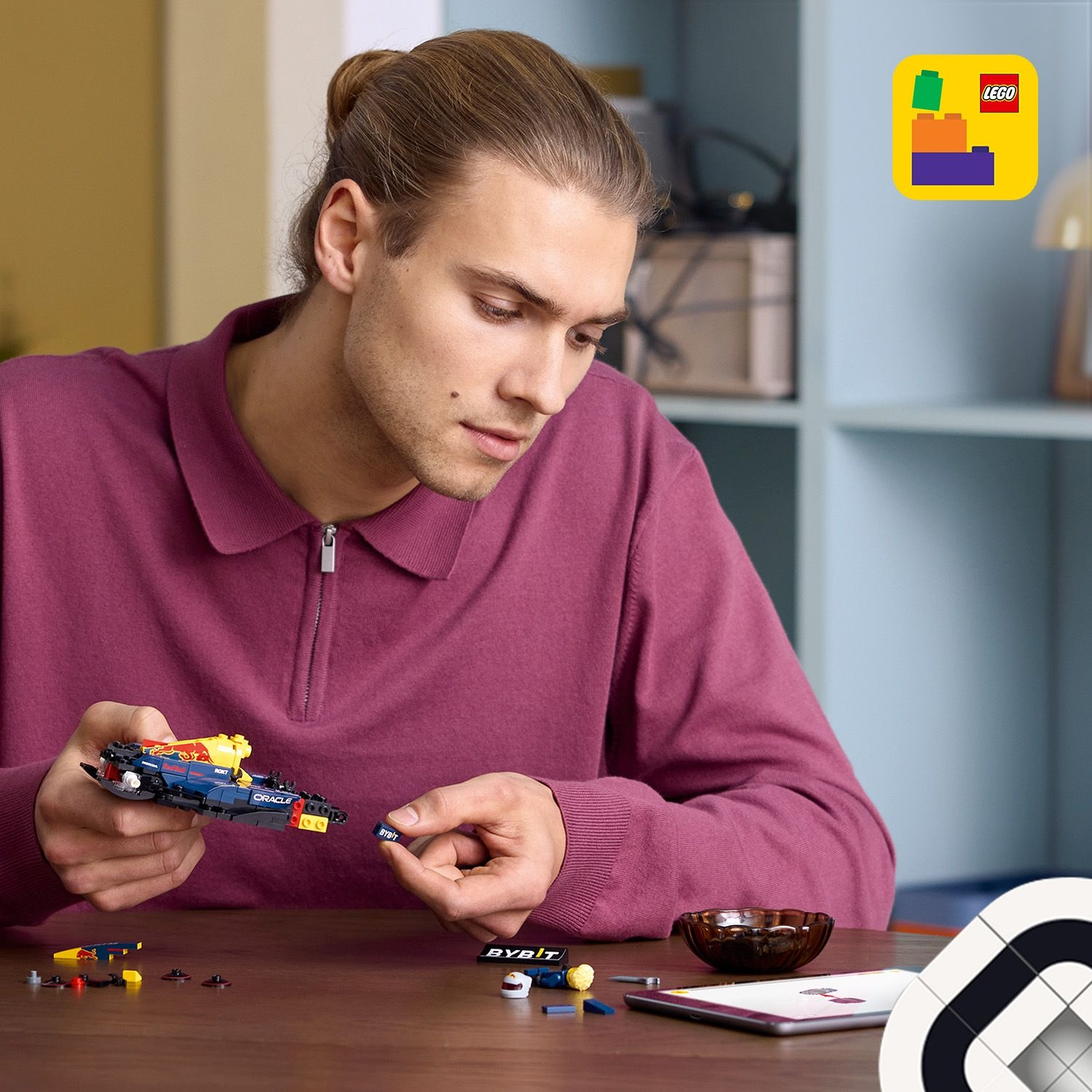 Un uomo assembla una macchina da corsa Lego. Indossa una camicia bordeaux e sullo sfondo ci sono un logo Lego e un tavolo.