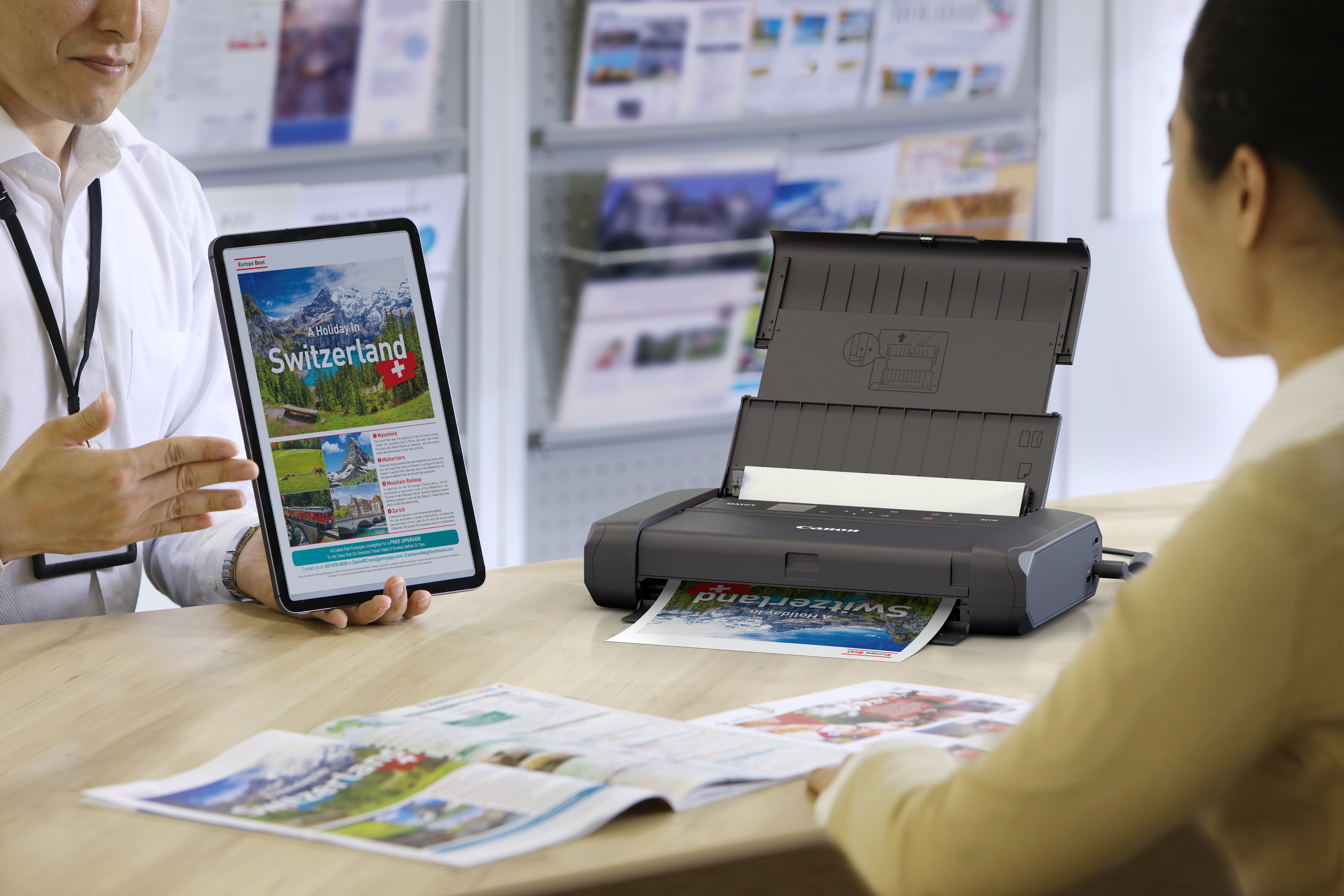 Un uomo mostra un tablet con una pubblicità sulla Svizzera. Una stampante stampa la stessa pubblicità. Una donna guarda la stampa.