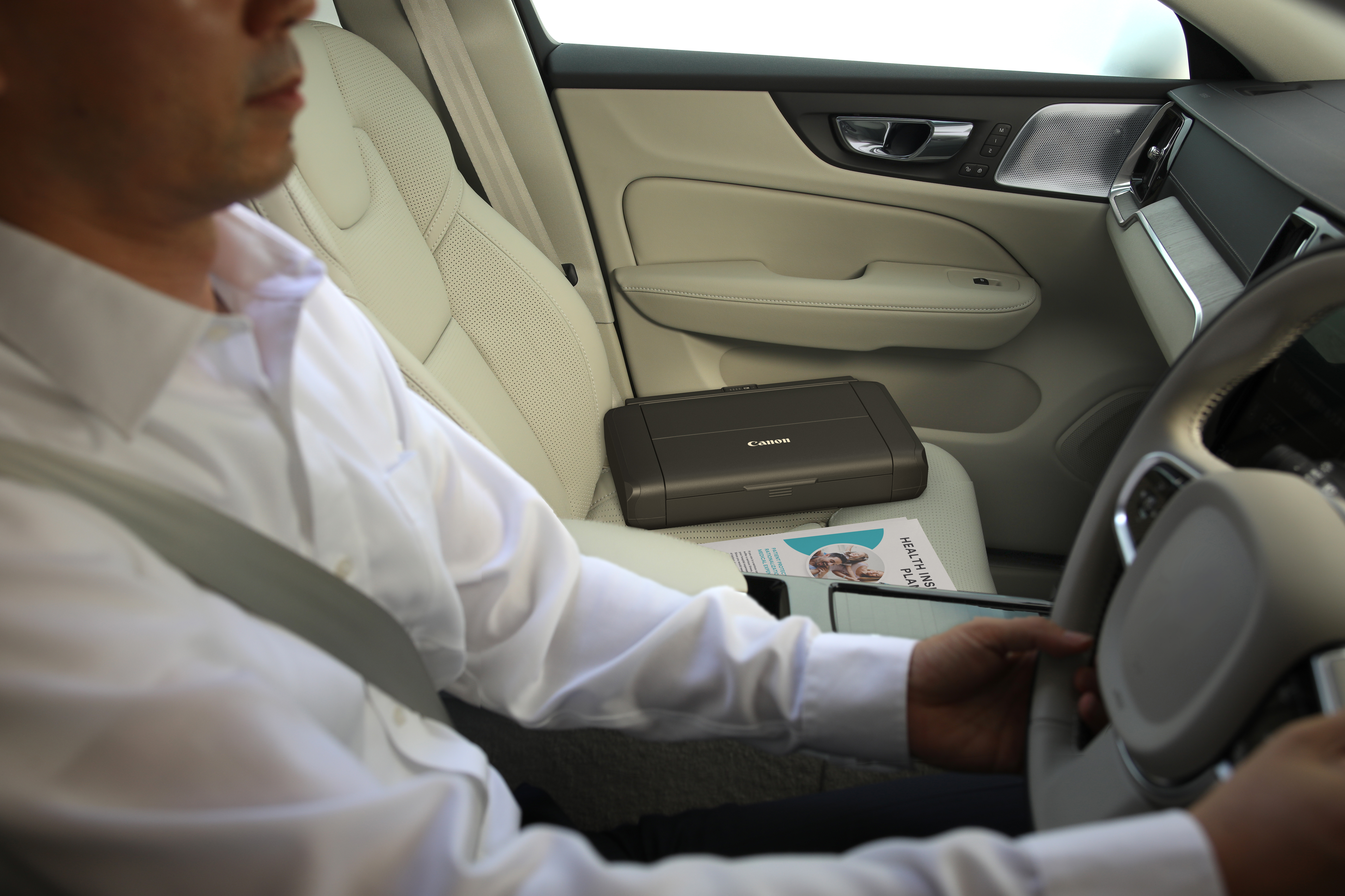 All'interno di un'auto, una persona con una camicia bianca tiene il volante. Una stampante Canon è sul sedile del passeggero.