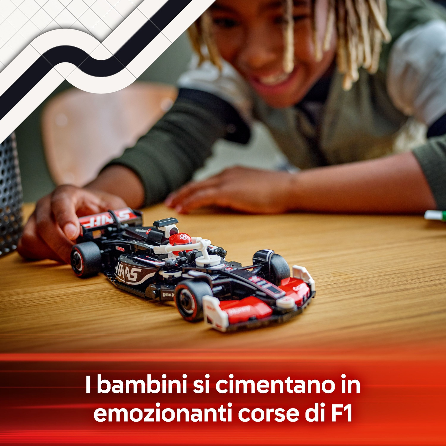 Un bambino sorride sopra un'auto da corsa F1 giocattolo su un tavolo di legno. L'auto è nera, rossa e bianca. Lo sfondo è sfocato.