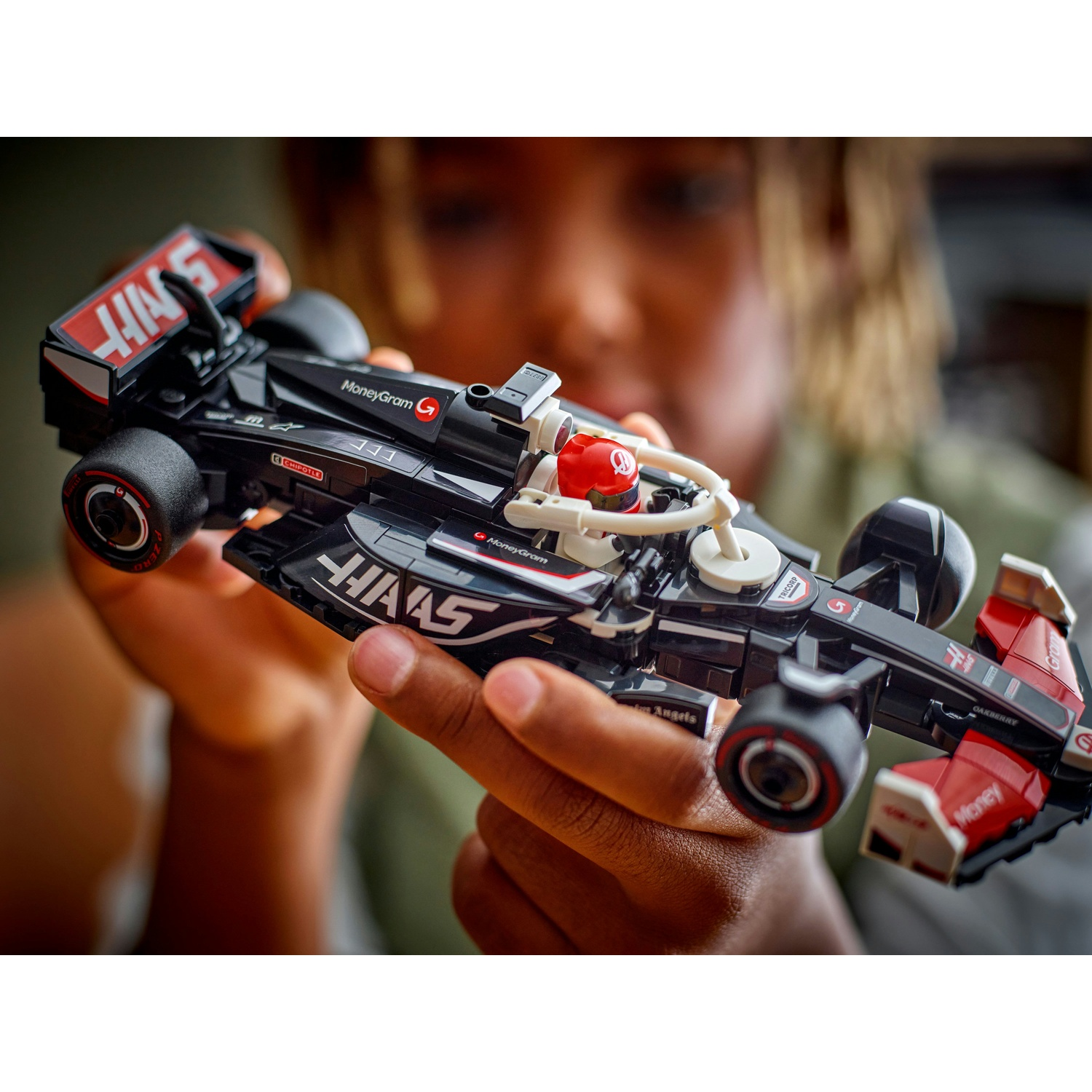 Una persona tiene in mano una macchina da corsa giocattolo nera e rossa. L'auto ha i loghi degli sponsor. La persona è fuori fuoco.