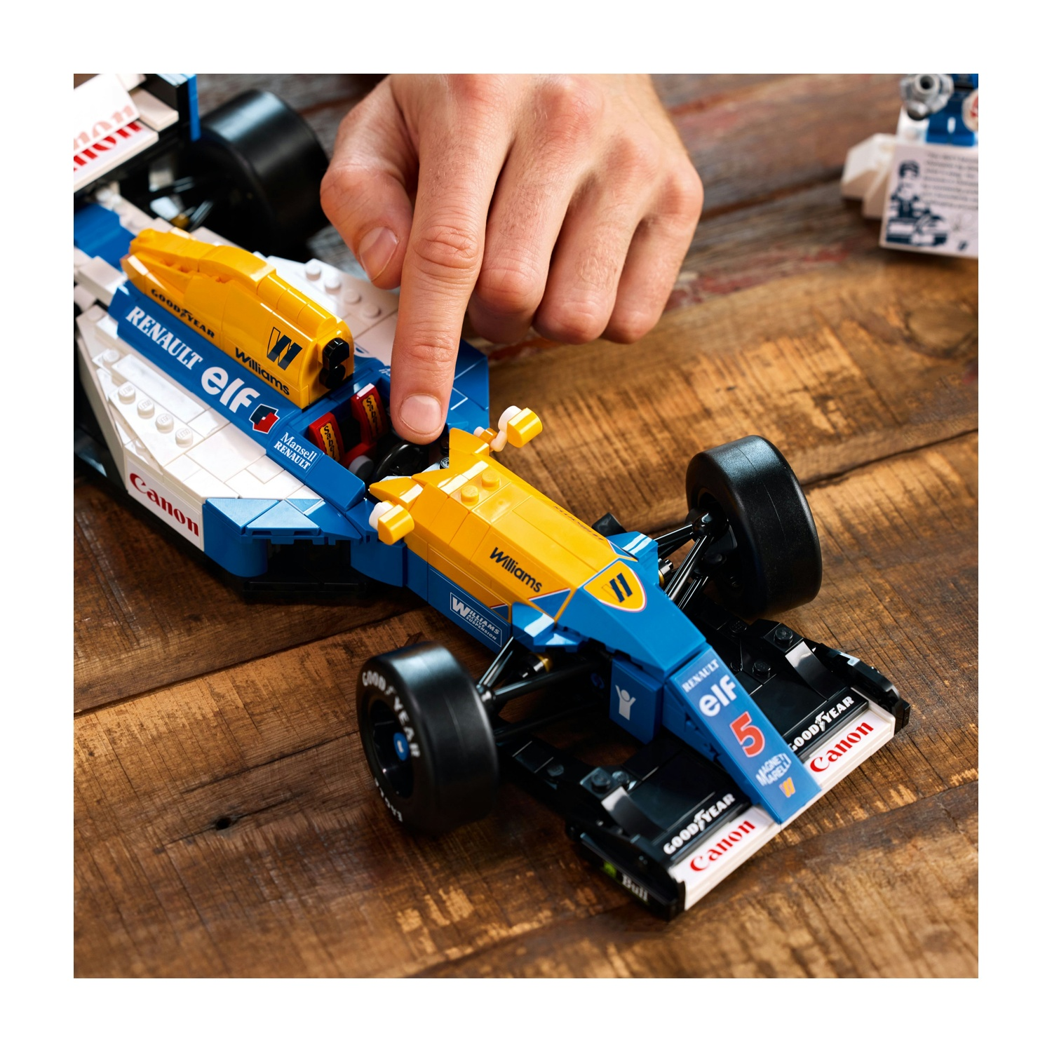 Una mano tocca un'auto da corsa Lego. L'auto è blu, gialla e bianca, con i loghi degli sponsor. È appoggiata su una superficie di legno.