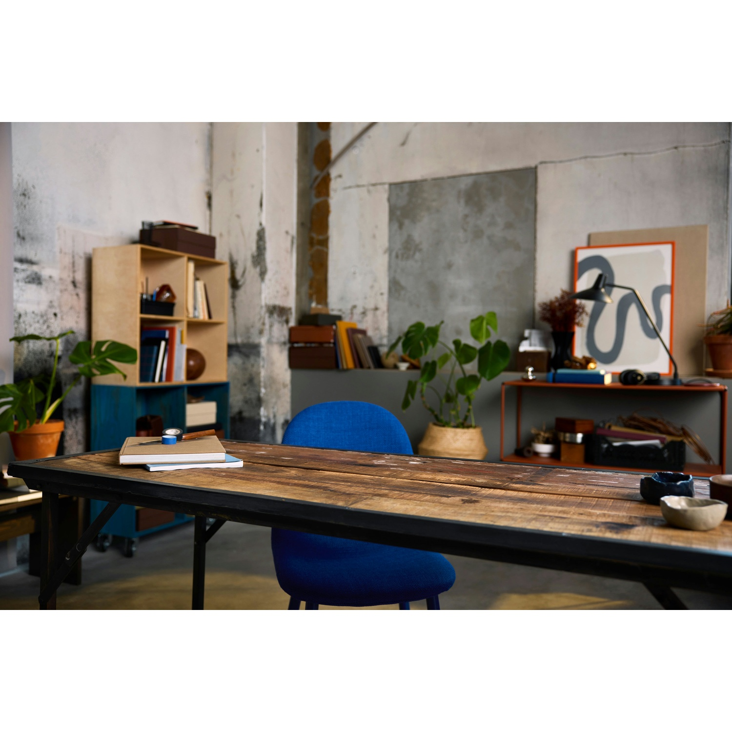 Un tavolo di legno con libri, una sedia blu e piante in una stanza con scaffali e una cornice.