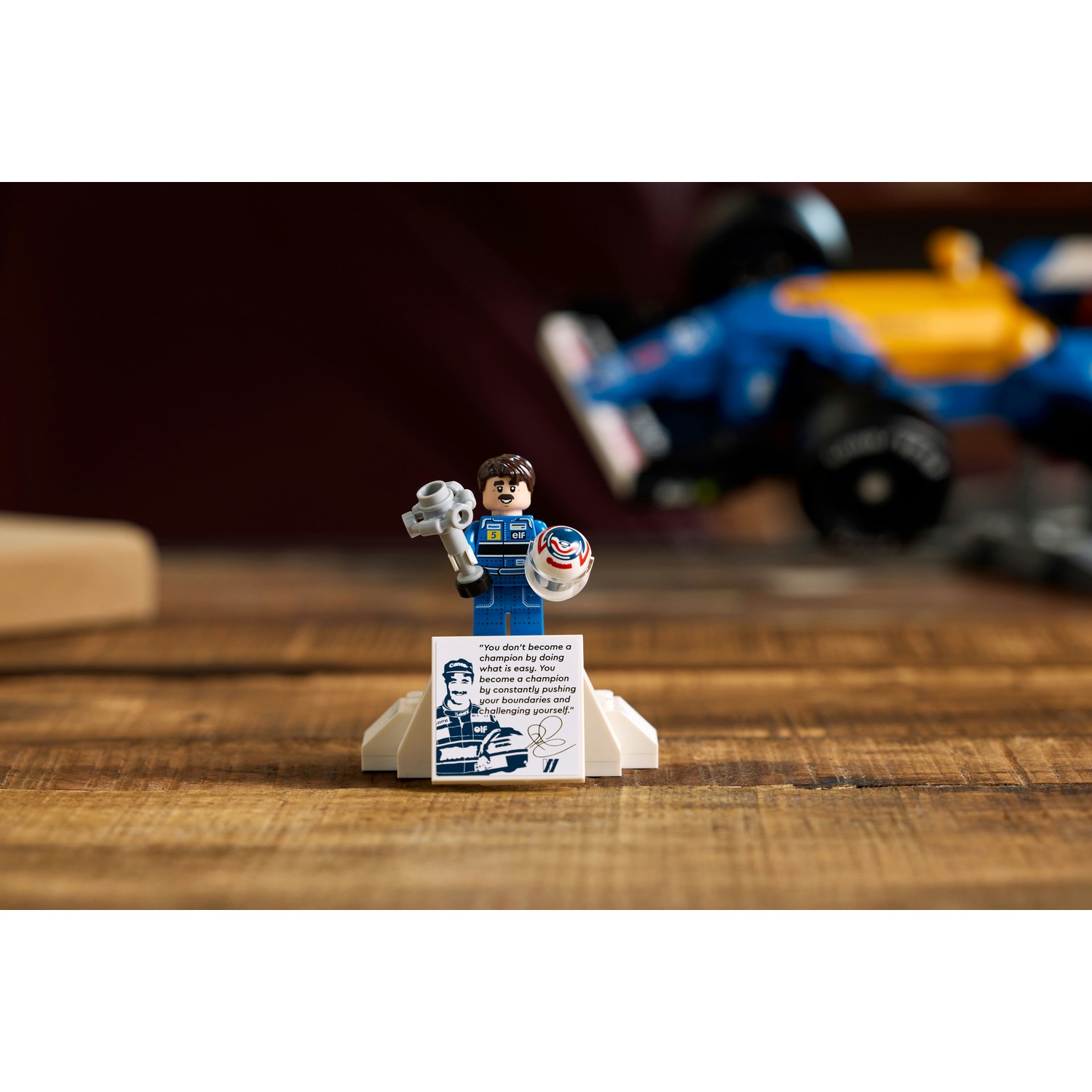 Un personaggio Lego tiene una chiave inglese e un casco, in piedi di fronte a un'auto da corsa blu e gialla. Un cartello con una citazione è davanti.