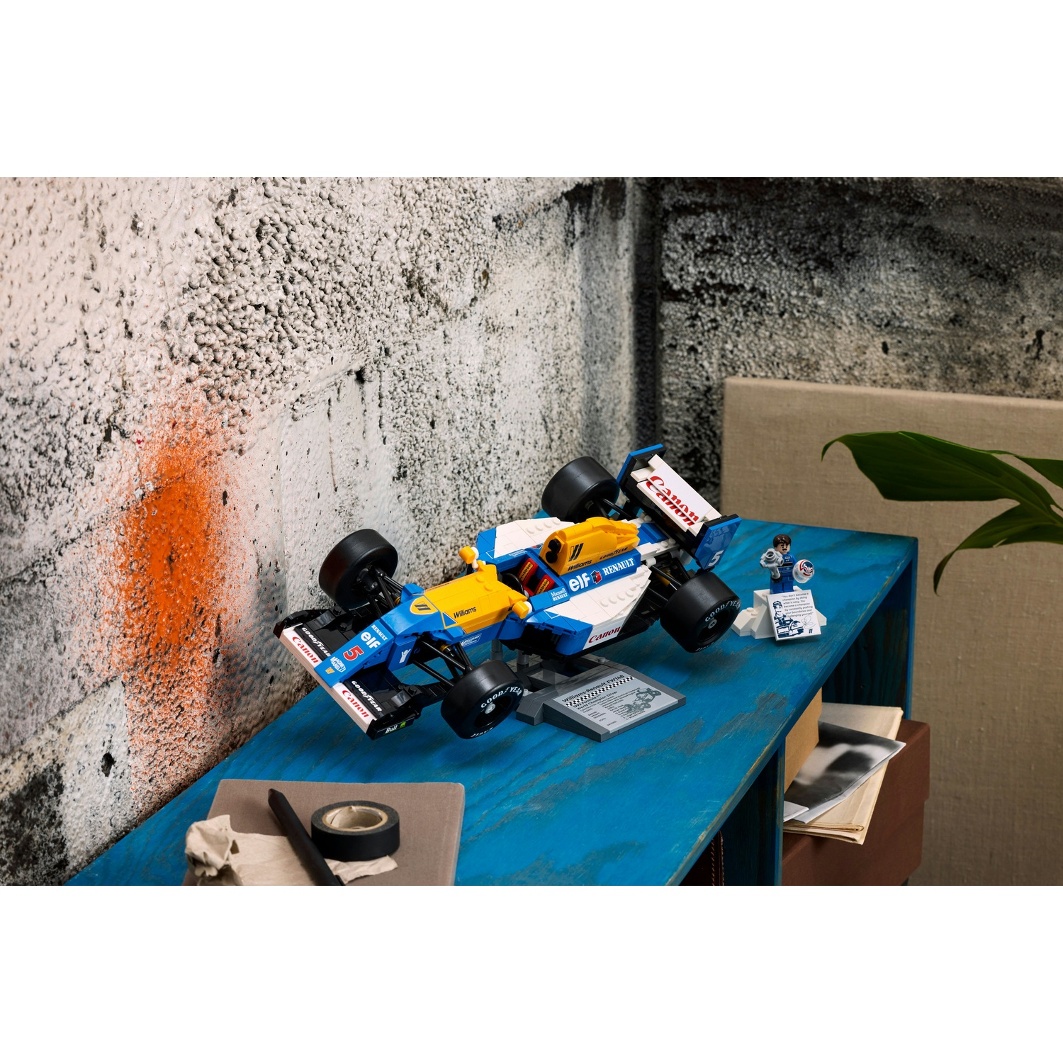 Un modello Lego di F1, blu e giallo, è su un tavolo blu. Lo sfondo è un muro strutturato con vernice arancione.