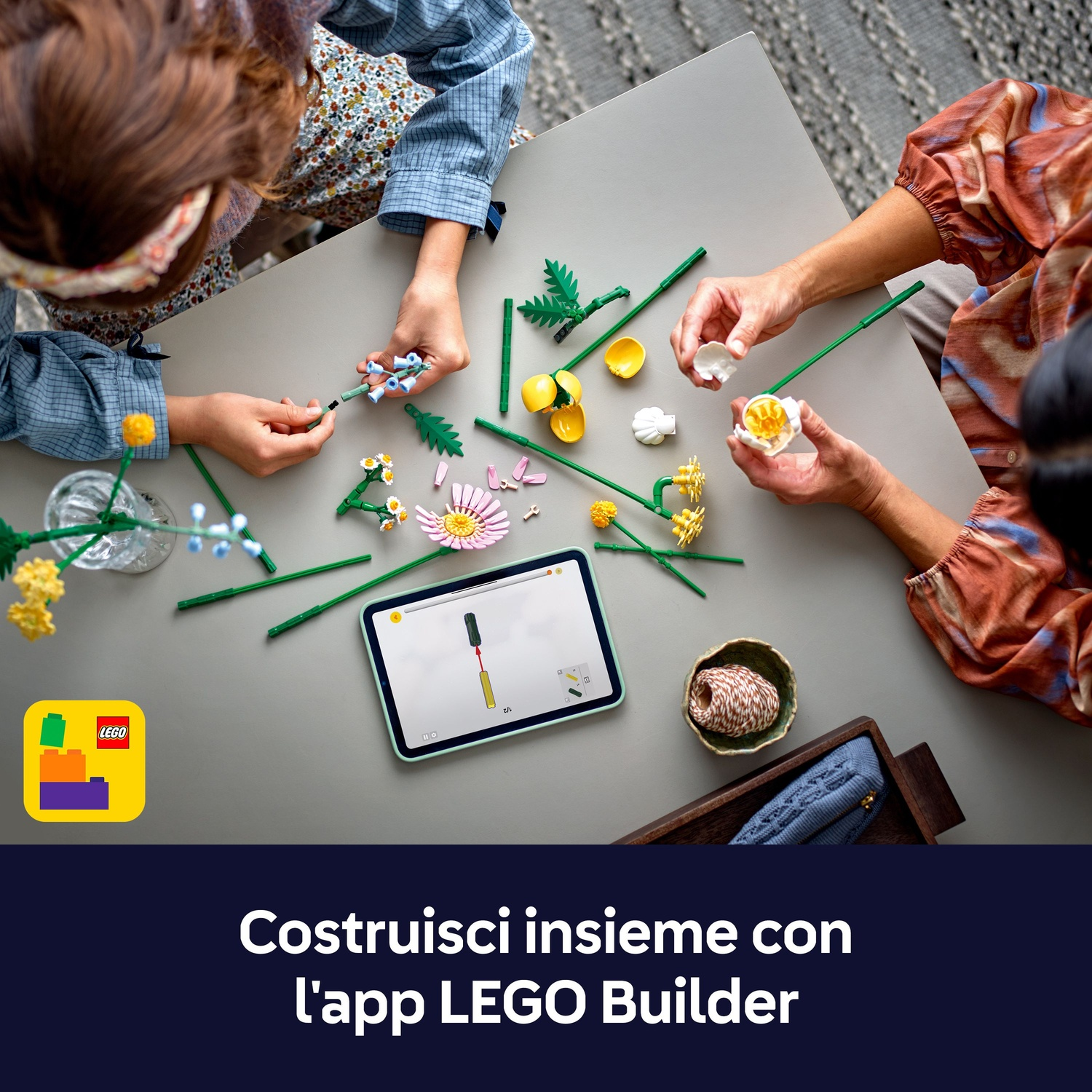 Due persone costruiscono fiori LEGO su un tavolo, usando un tablet. Logo LEGO visibile. Varie parti di fiori sono presenti.