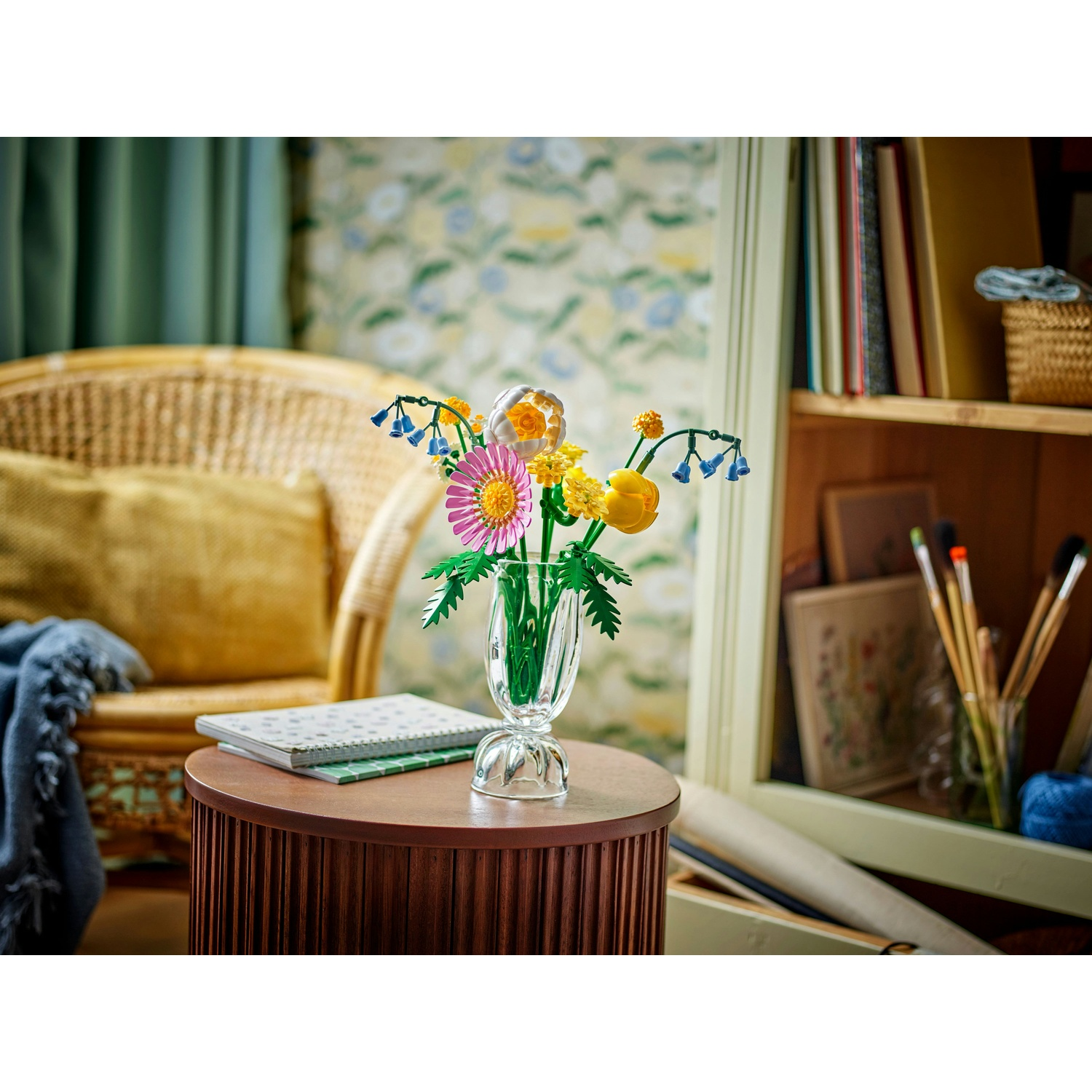 Un vaso trasparente con fiori colorati su un tavolo marrone. Una sedia, libri e opere d'arte sullo sfondo sfocato.