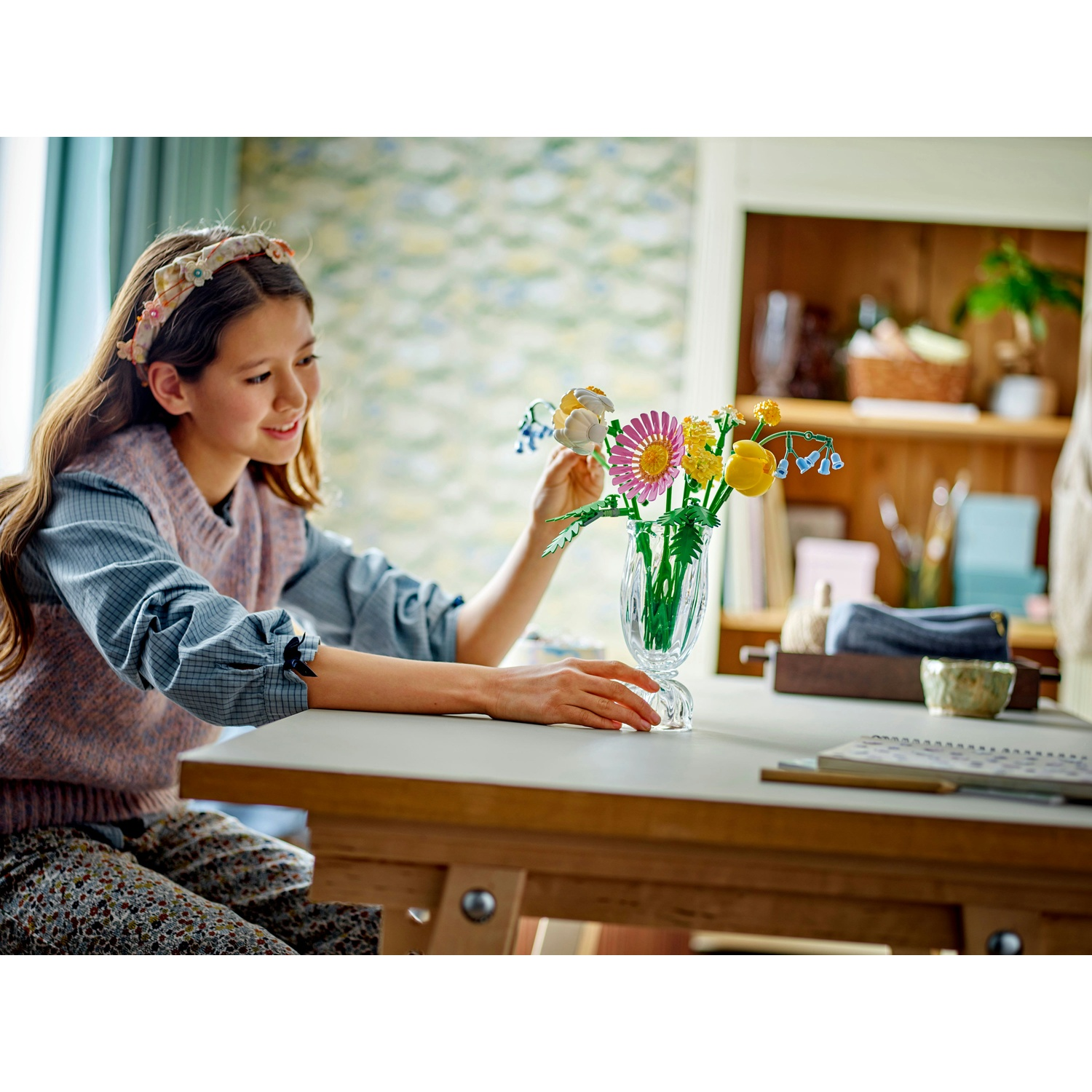 Una ragazza sorride a un vaso con fiori colorati. Indossa una fascia floreale e un maglione. Un tavolo di legno è davanti.