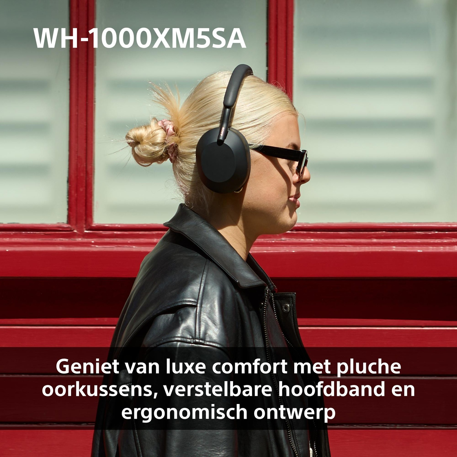 Een vrouw draagt een zwarte koptelefoon en zonnebril. Ze heeft blond haar en een zwarte leren jas. Een rode muur is op de achtergrond.