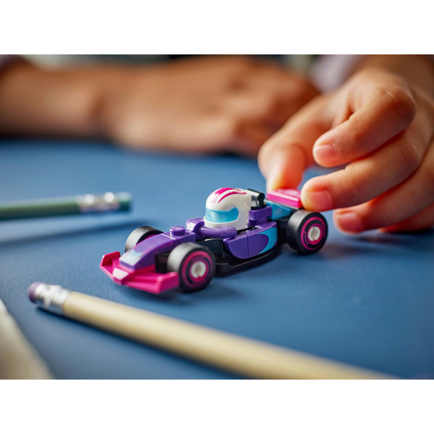 La mano di un bambino tocca un'auto da corsa giocattolo viola e rosa. Matite in primo piano, con una superficie blu sullo sfondo.
