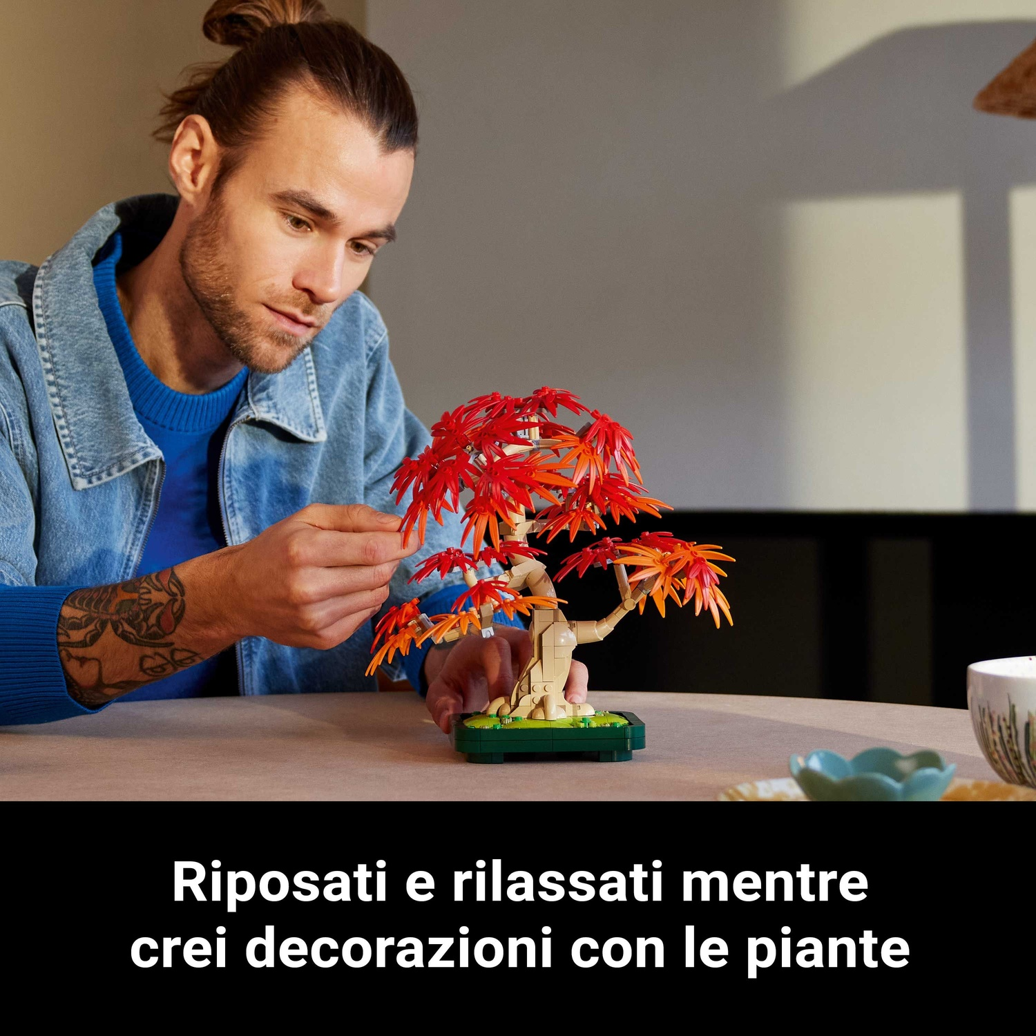 Un uomo con uno chignon guarda un bonsai. L'albero ha foglie rosse e arancioni. Ci sono alcuni oggetti sul tavolo.