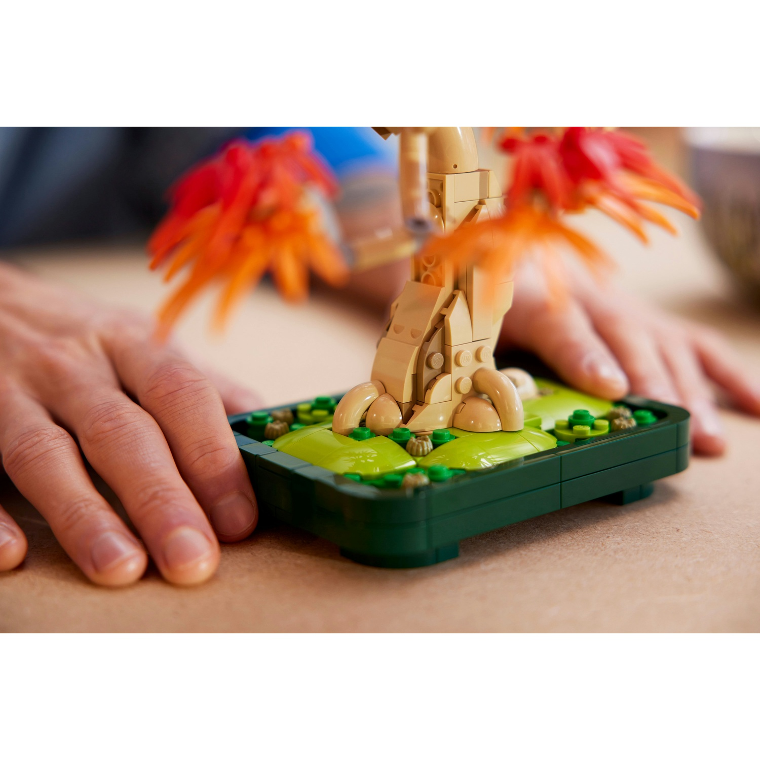 Un modello di albero LEGO con foglie arancioni e tronco marrone chiaro è in un contenitore verde. Le mani sono sul tavolo accanto al modello.
