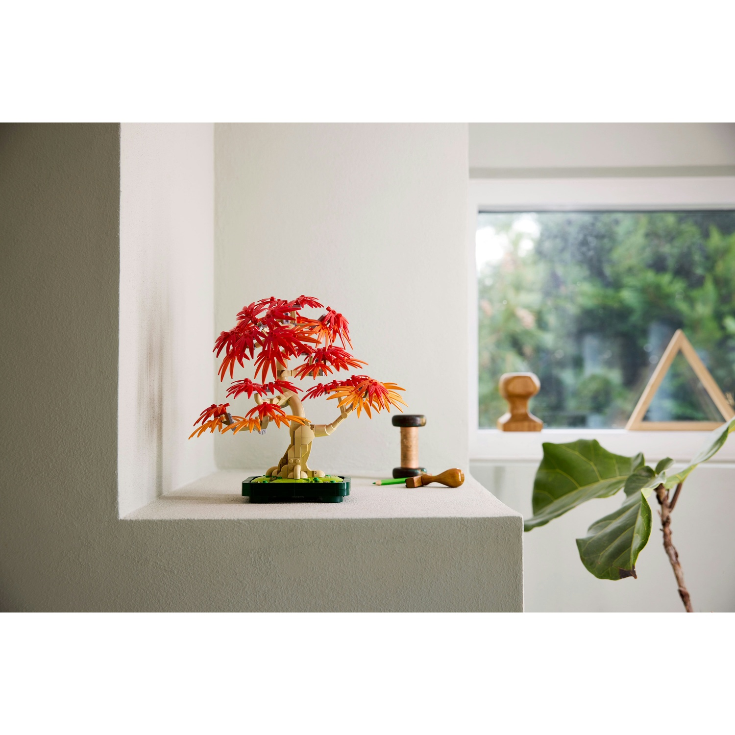 Un bonsai Lego con foglie rosse e arancioni è su una sporgenza bianca. Sullo sfondo una finestra e piante. Sono presenti anche accessori.