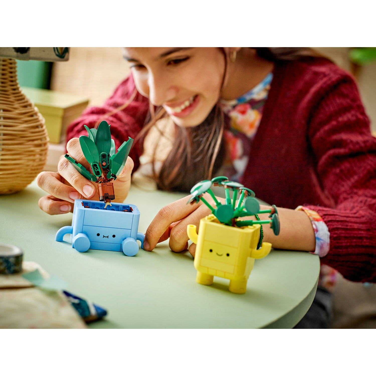 Una ragazza sorridente gioca con le piante Lego. Ci sono due fioriere Lego, una blu e una gialla, su un tavolo verde.