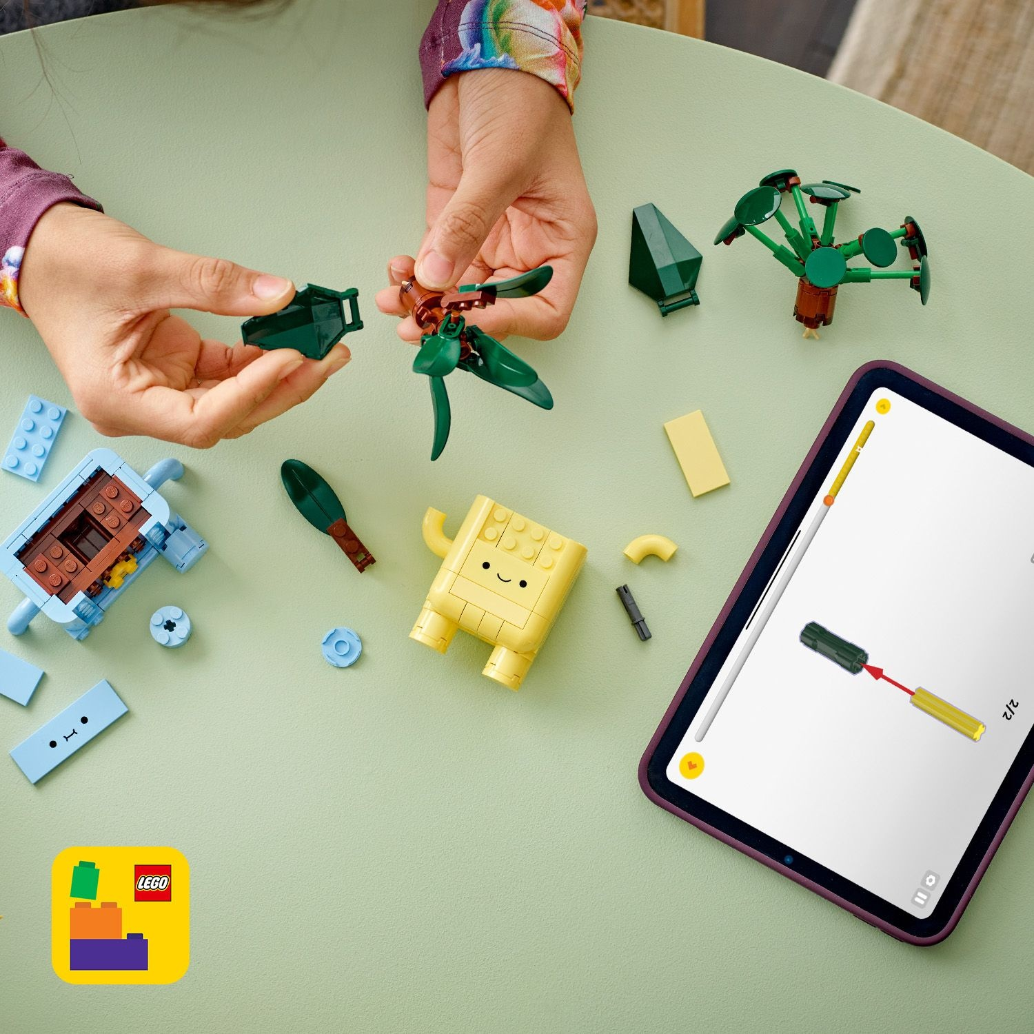 Le mani assemblano pezzi Lego su un tavolo verde. Ci sono vari elementi Lego e un tablet che mostra le istruzioni. Un logo Lego è visibile.