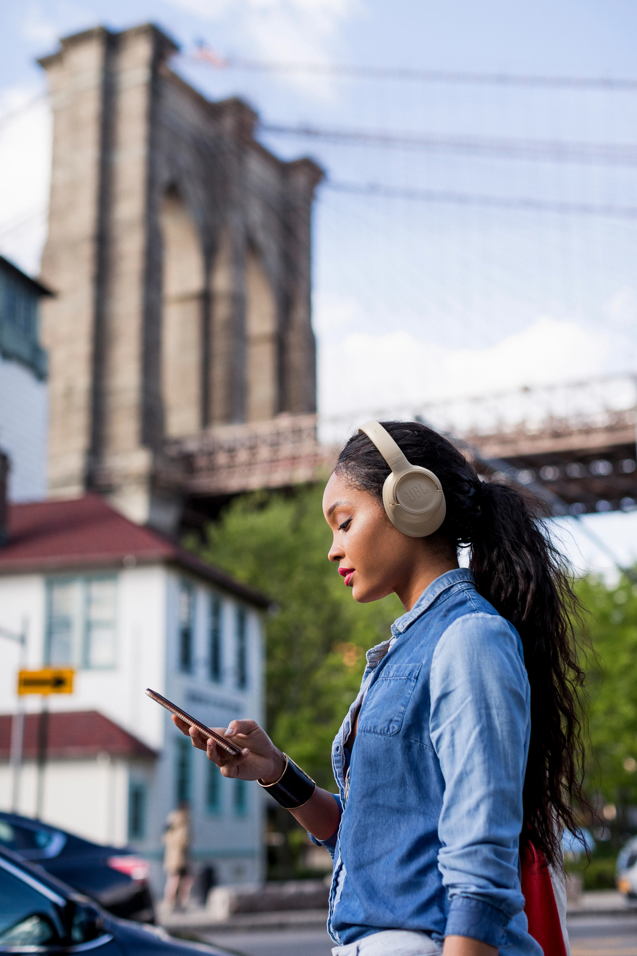 Frau mit Kopfhörern, Jeanshemd, Handy. Brooklyn Bridge im Hintergrund. Gebäude, Autos und Bäume sind ebenfalls sichtbar.