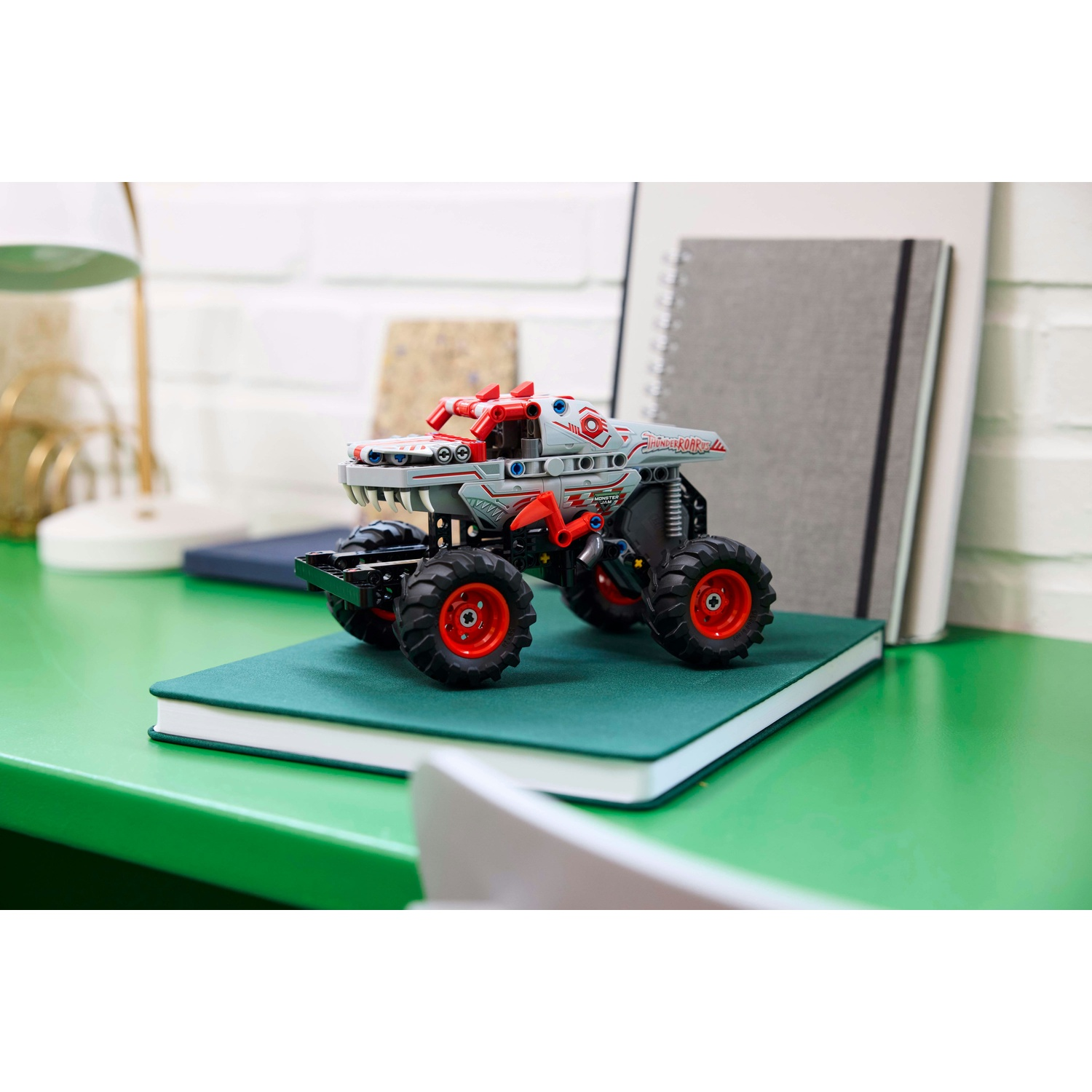 Un monster truck giocattolo è su un quaderno verde. Ha dettagli rossi e grigi e pneumatici neri. Lo sfondo mostra quaderni e una lampada.