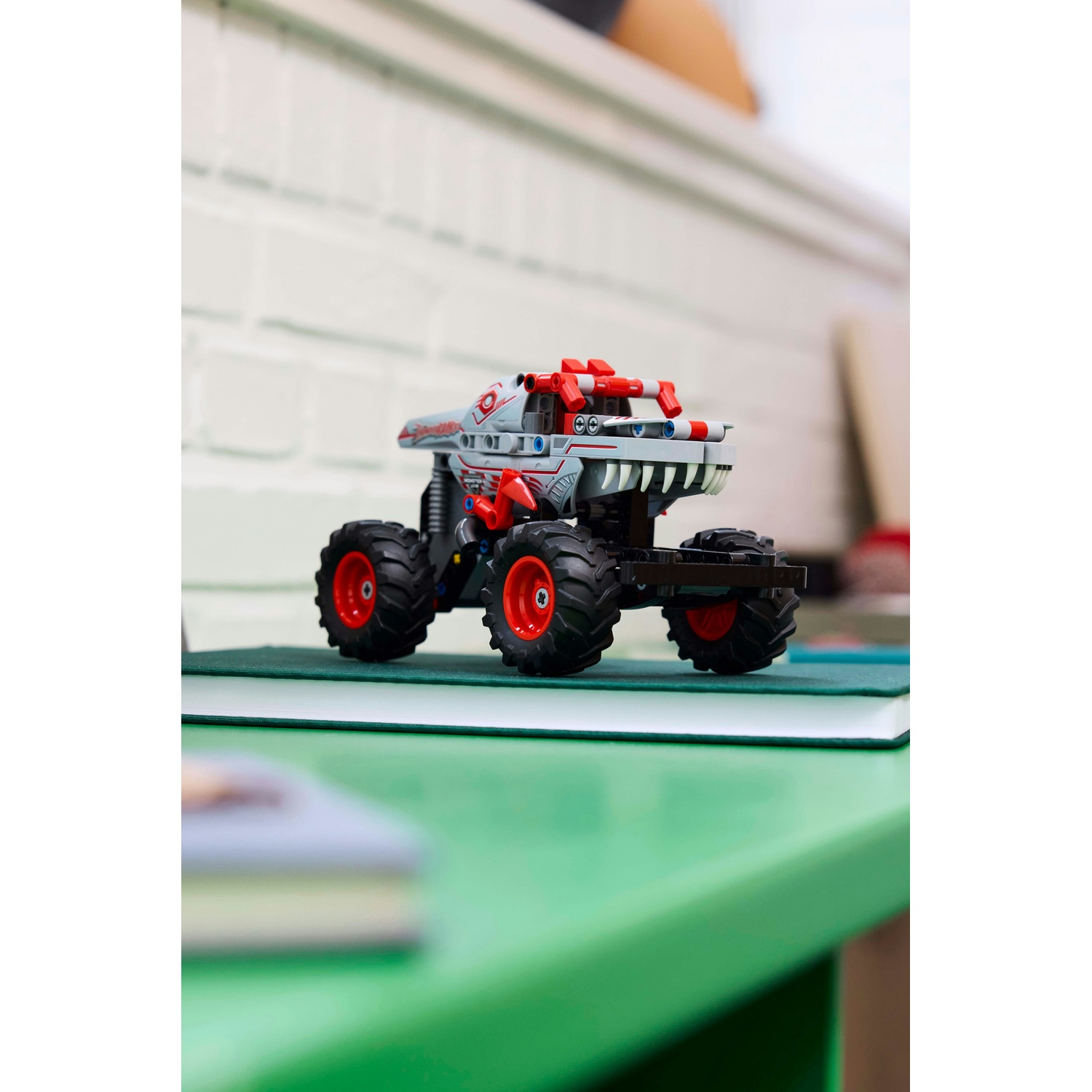 Un monster truck giocattolo con carrozzeria grigia, dettagli rossi e ruote nere su una superficie verde con sfondo sfocato.