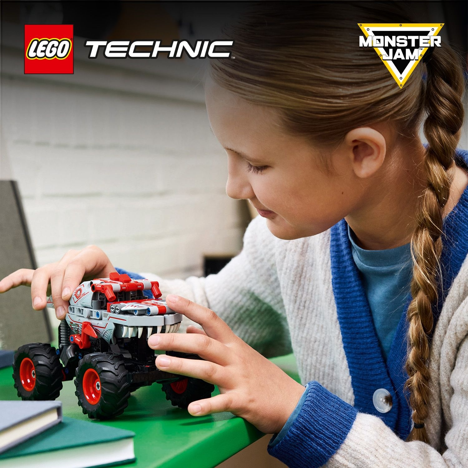 Una ragazza gioca con un camion LEGO Technic Monster Jam su un tavolo verde. Il camion è bianco, rosso e nero. C'è un libro vicino.