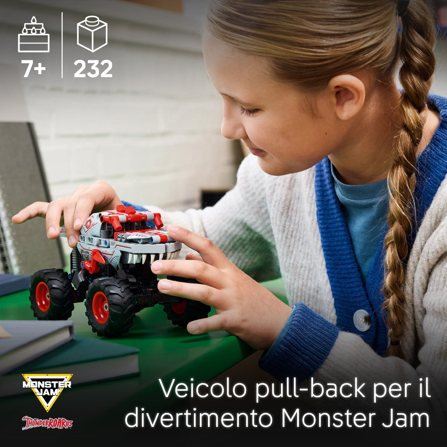 Una ragazza gioca con un camion giocattolo Monster Jam. Il giocattolo è grigio, rosso e bianco. Lo sfondo presenta libri e una superficie verde.