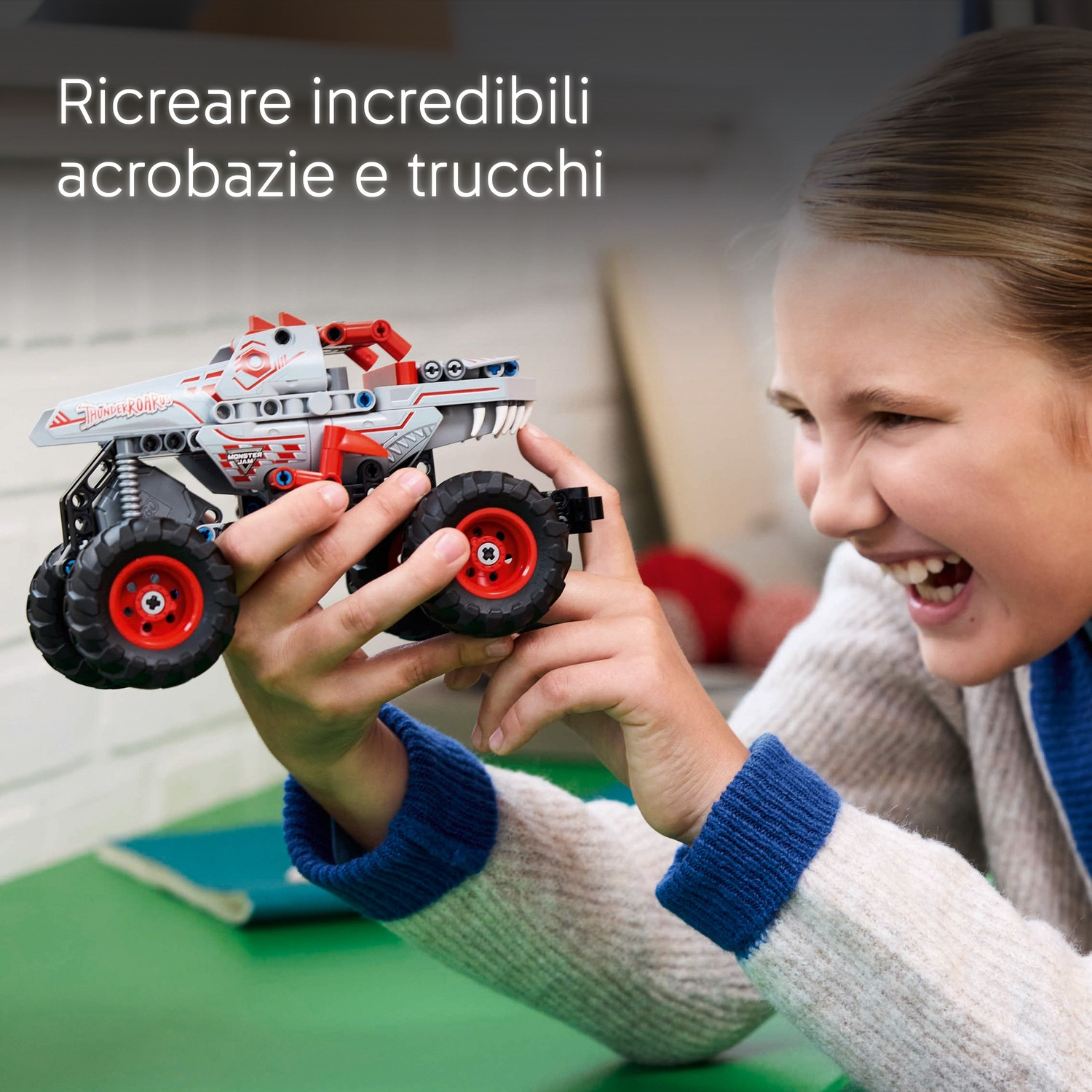 Una ragazza sorride mentre tiene in mano un monster truck giocattolo. Il camion è grigio, rosso e nero, con ruote grandi. Lo sfondo è una superficie verde.